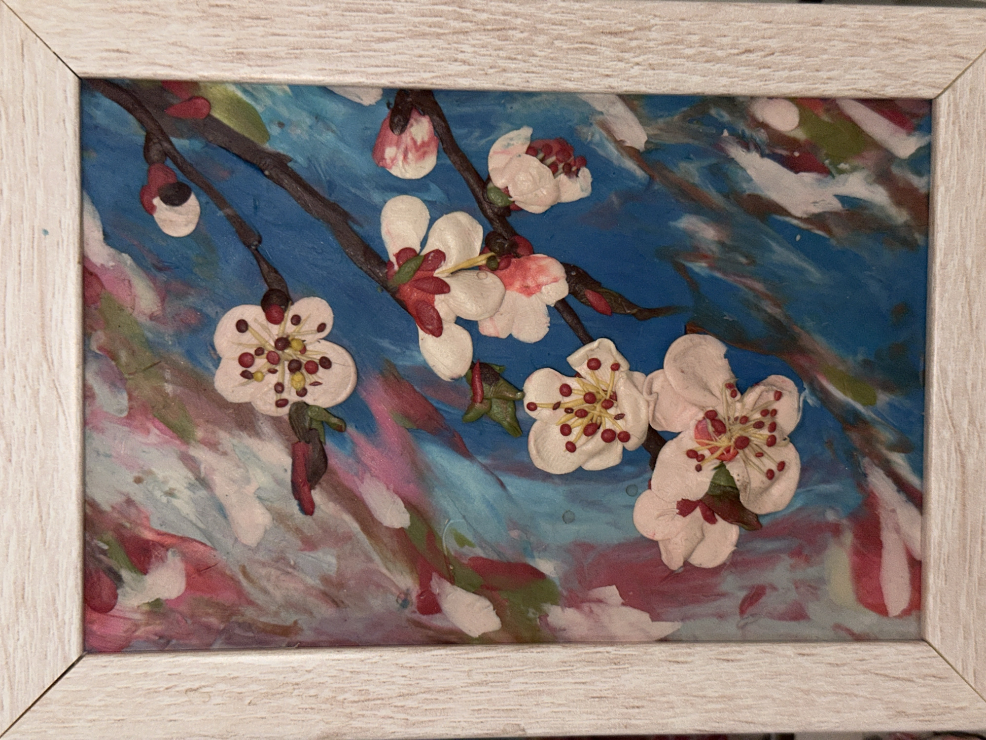 A framed artwork featuring blossom branch motifs. The background consists of broad strokes of blue, white, and pink. The blossoms and branch are sculpted with clay, creating a three-dimensional effect. The petals are white with pink tinges, and the centers are adorned with red and yellow details. The frame is light-colored wood. The artwork is located in Unknown, Unknown.