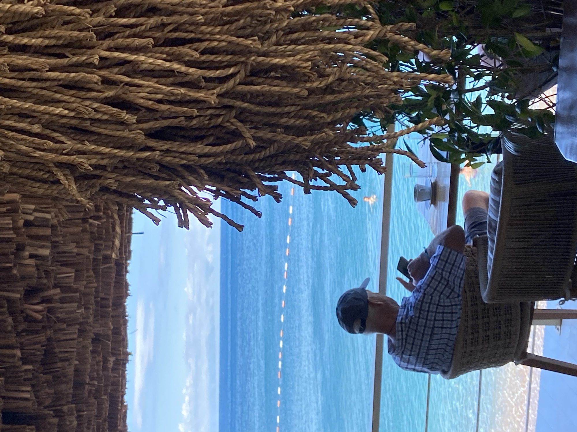 This image depicts a man sitting in a woven chair at an outdoor establishment, likely a cafe or restaurant, overlooking the Adriatic Sea in Petrovac centar, Montenegro. The man, viewed from the back, wears a checkered shirt and a cap, and is engrossed in his mobile phone. In front of him, on a wooden table, is a cup of coffee and a glass of water. The foreground is partially obscured by hanging ropes and greenery, suggesting a rustic or bohemian decor. The background features the clear blue sea, with a line of buoys marking a swimming area. A person is visible swimming in the water. The sky is bright blue with scattered clouds, indicating a pleasant day. The architecture of the establishment is characterized by a thatched roof and woven furniture, adding to the relaxed, seaside ambiance.