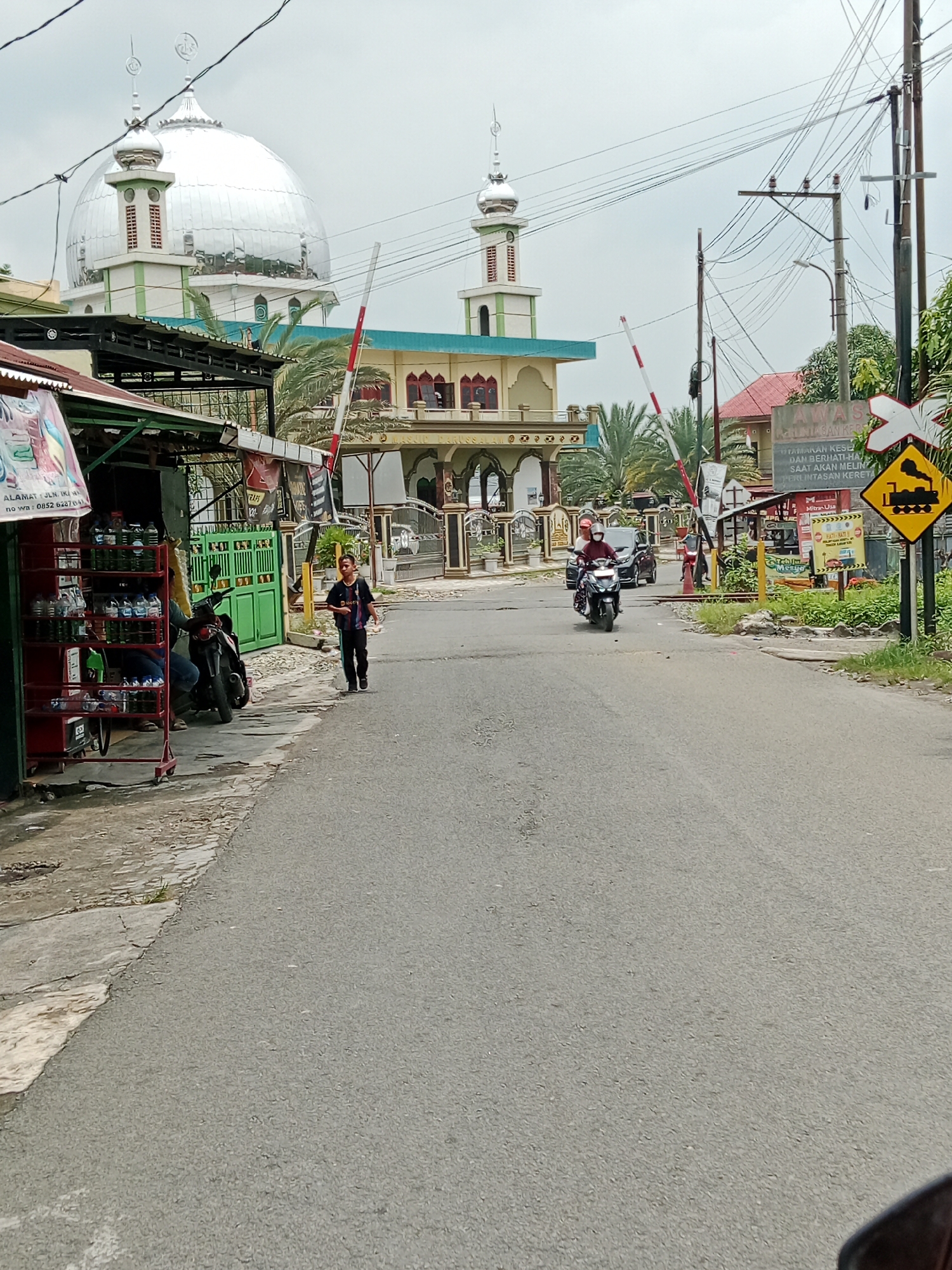 The image depicts a street scene in the City of Binjai, Indonesia, near a railway crossing and a mosque. The foreground shows a paved road with a slight curve. On the left side of the road, there is a small roadside stall with shelves displaying various bottled drinks. A motorcycle is parked near the stall, and a person is partially visible sitting on it. A young man in dark clothing is walking on the sidewalk next to the stall, facing away from the camera.
In the mid-ground, a railway crossing with striped barrier arms is visible, one of which is lowered. A motorcycle with two riders, wearing helmets, is approaching the crossing from the distance. Behind the crossing, a building with a large silver dome and minarets, identified as a mosque, is prominent. The mosque has light-colored walls with green accents and arched windows. Further down the road, a yellow diamond-shaped sign with a black train silhouette indicates a railway crossing. Utility poles with numerous wires are also present. The background consists of trees and buildings under an overcast sky. The overall scene suggests a typical day in a residential or commercial area of the city with active traffic and pedestrian movement.