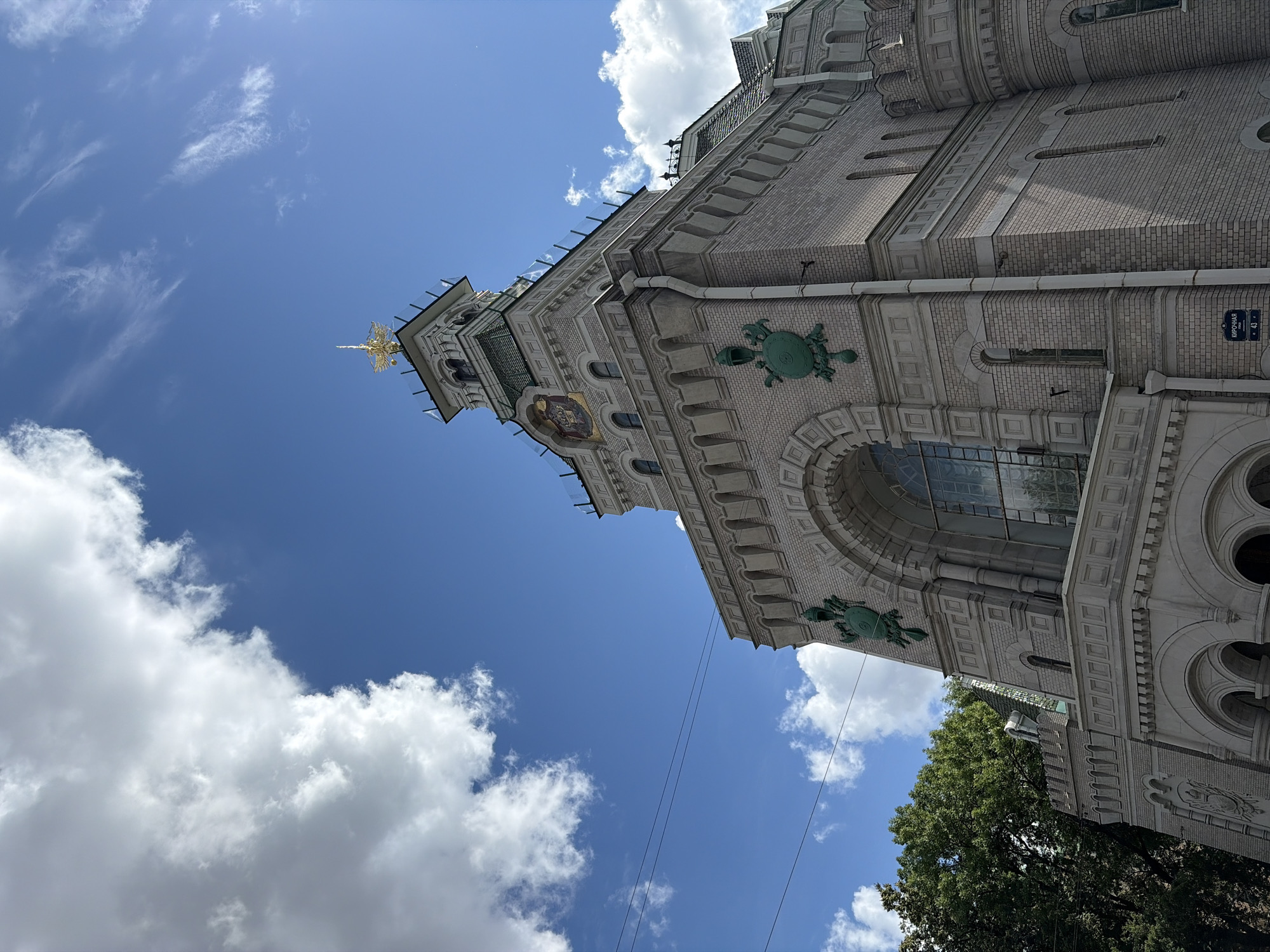 The image captures a low-angle view of a light-colored brick building with a prominent tower against a bright blue sky dotted with white clouds. The building features intricate architectural details, including arched windows, decorative green crests resembling shields with helmets, and a golden spire atop the tower. A dark green tree is visible to the left of the building, and power lines stretch across the sky. A street sign with Russian text and the number "43" is affixed to the building's lower section. The architecture suggests a historical or ecclesiastical structure, and the overall impression is one of grandeur and history. Based on the street sign, the location is likely Kirochnaya Ulitsa, Saint Petersburg, Russia.