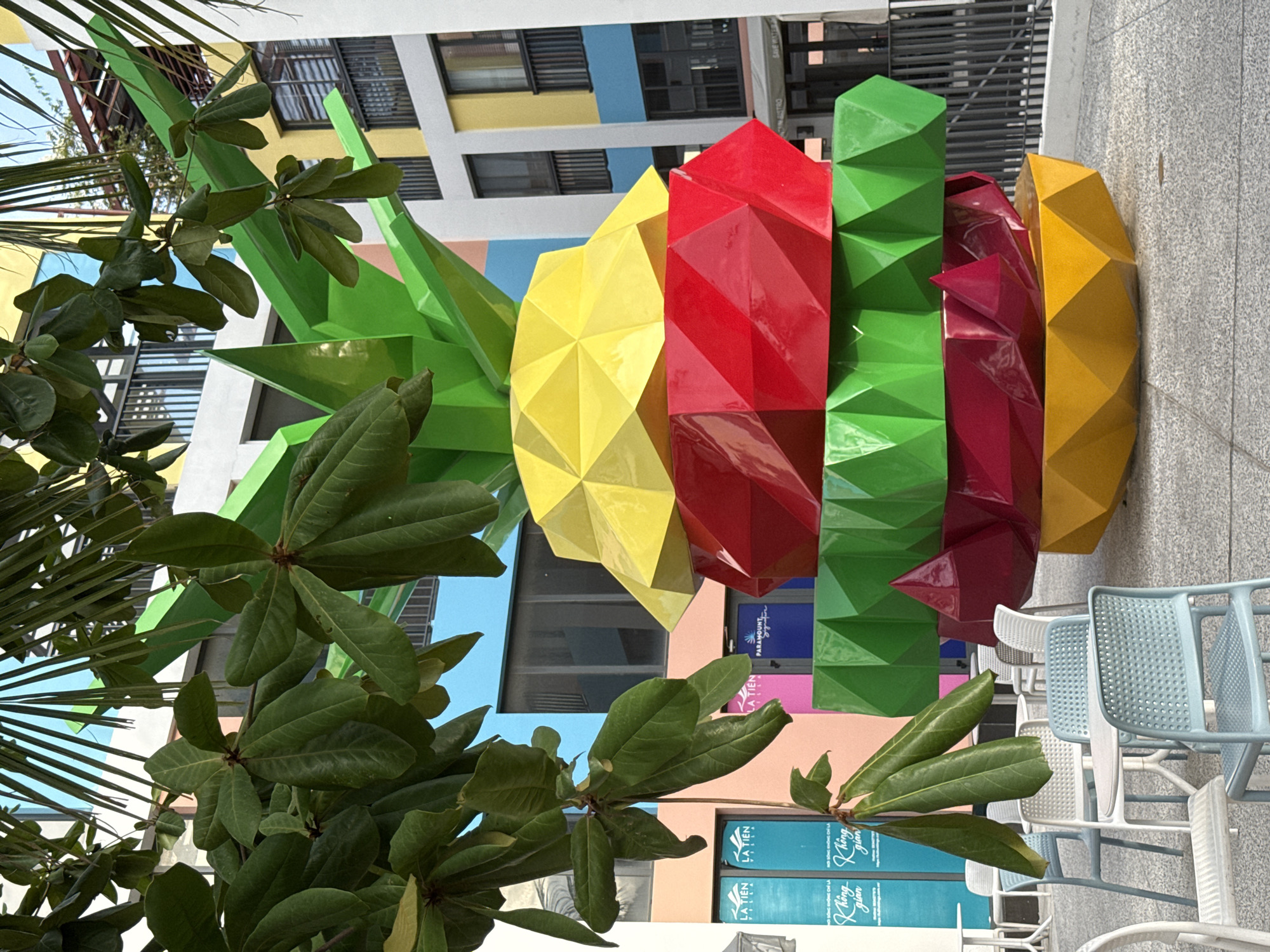 In Nha Trang, Vietnam, a large, multi-colored abstract sculpture resembling a pineapple or burger is displayed outdoors in a paved area with tables and chairs. The sculpture is made of geometric, faceted segments in shades of green, yellow, red, and pink. It is partially obscured by lush green foliage from overhanging trees. In the background, a modern building with brightly colored panels in yellow, blue, pink, and white is visible, featuring balconies and windows. Several cafe tables and chairs, some in light blue and white plastic, are arranged around the sculpture. Doors with signage, including "LA TIEN VILLA" and "PARAMOUNT," are present on the building's lower level. The scene appears to be a public outdoor space, possibly part of a resort or commercial area, designed for relaxation and visual appeal. There are no people visible in the image.