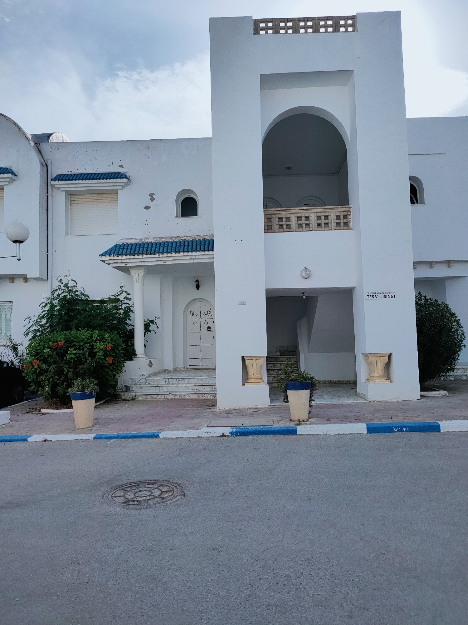 The image shows a white building in Mrezga, Tunisia, with blue accents on the roof tiles and a blue and white painted curb. The building features an arched entryway on the upper level and a covered entrance on the lower level with a white door. There are stairs leading to the lower entrance, and potted plants are placed on either side of the entryway. A street with a manhole cover is visible in the foreground. The sky is overcast.