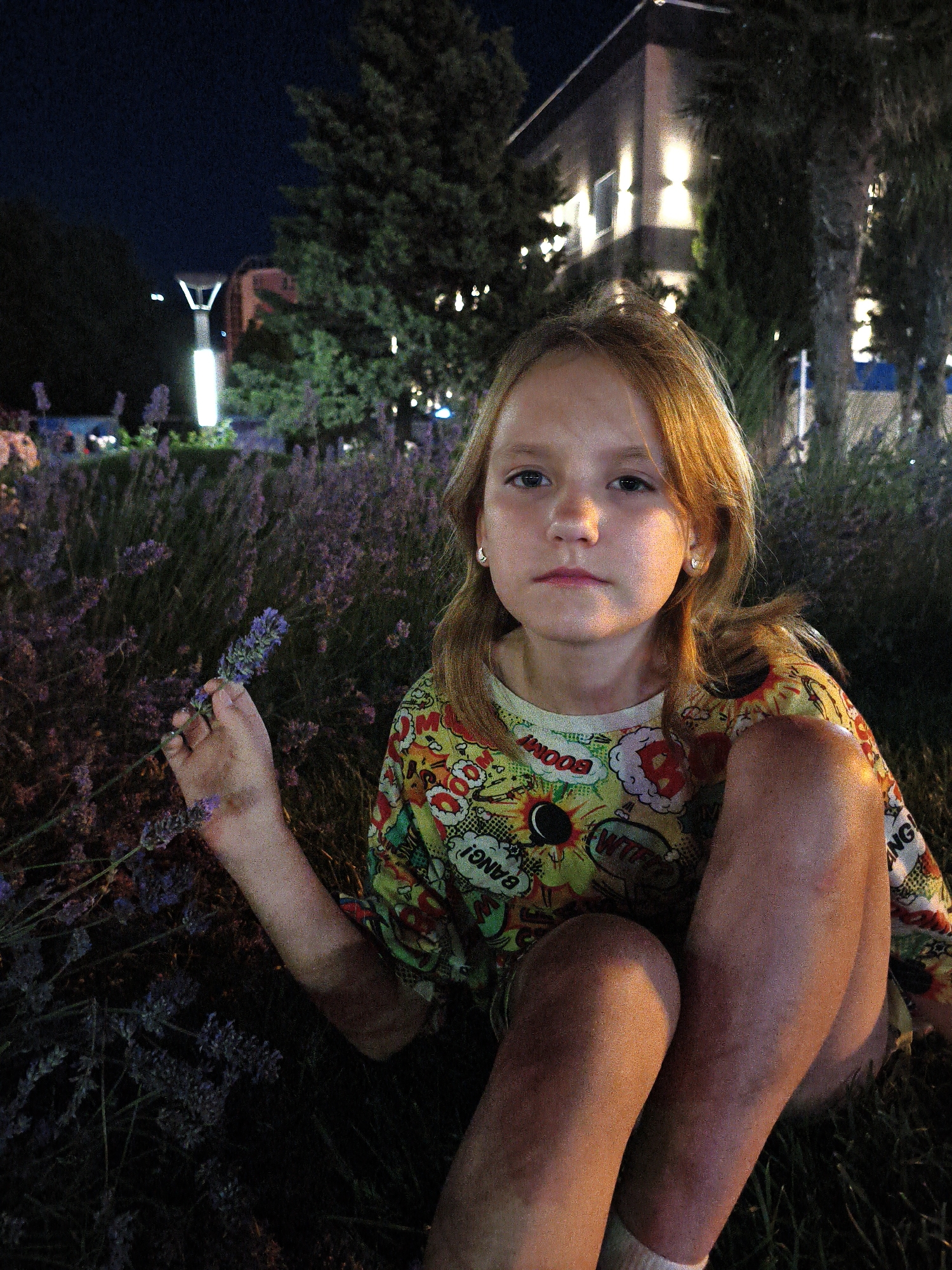 The image features a young girl sitting in a field of lavender at night. The girl has fair skin, blonde hair, and is looking directly at the camera with a neutral expression. She is wearing a colorful t-shirt with comic book-style graphics and the words "BOOM!", "WHIZ!", and "BANG!". Her right hand is holding a sprig of lavender. In the background, a modern building with illuminated windows is visible, surrounded by trees. A tall lamp post with a V-shaped top and a bright light is also present. The scene appears to be set in an outdoor urban or suburban area, given the presence of the building and lamp post, and the lavender suggests a park or garden setting. The overall lighting suggests it is nighttime, with artificial lights illuminating the scene. The presence of lavender indicates a temperate climate. The context provided states the location is Krinitsa, Russia.