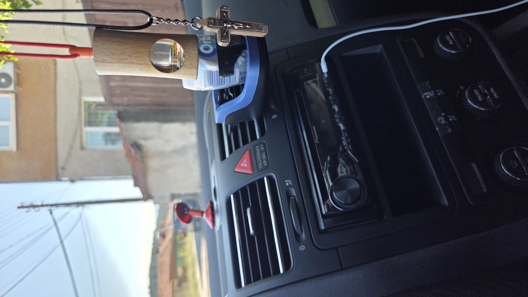 The image shows the interior of a car, looking out through the windshield. The dashboard is visible in the foreground, featuring air vents, a radio, and various controls. Hanging from the rearview mirror is a wooden car air freshener with a small glass vial containing a liquid, and a rosary with a crucifix. Also attached to the air vent is a blue plastic holder containing a pack of Orbit gum. Outside the car, a building with a red-tiled roof and a wooden fence can be seen. The sky is bright and appears to be daytime.