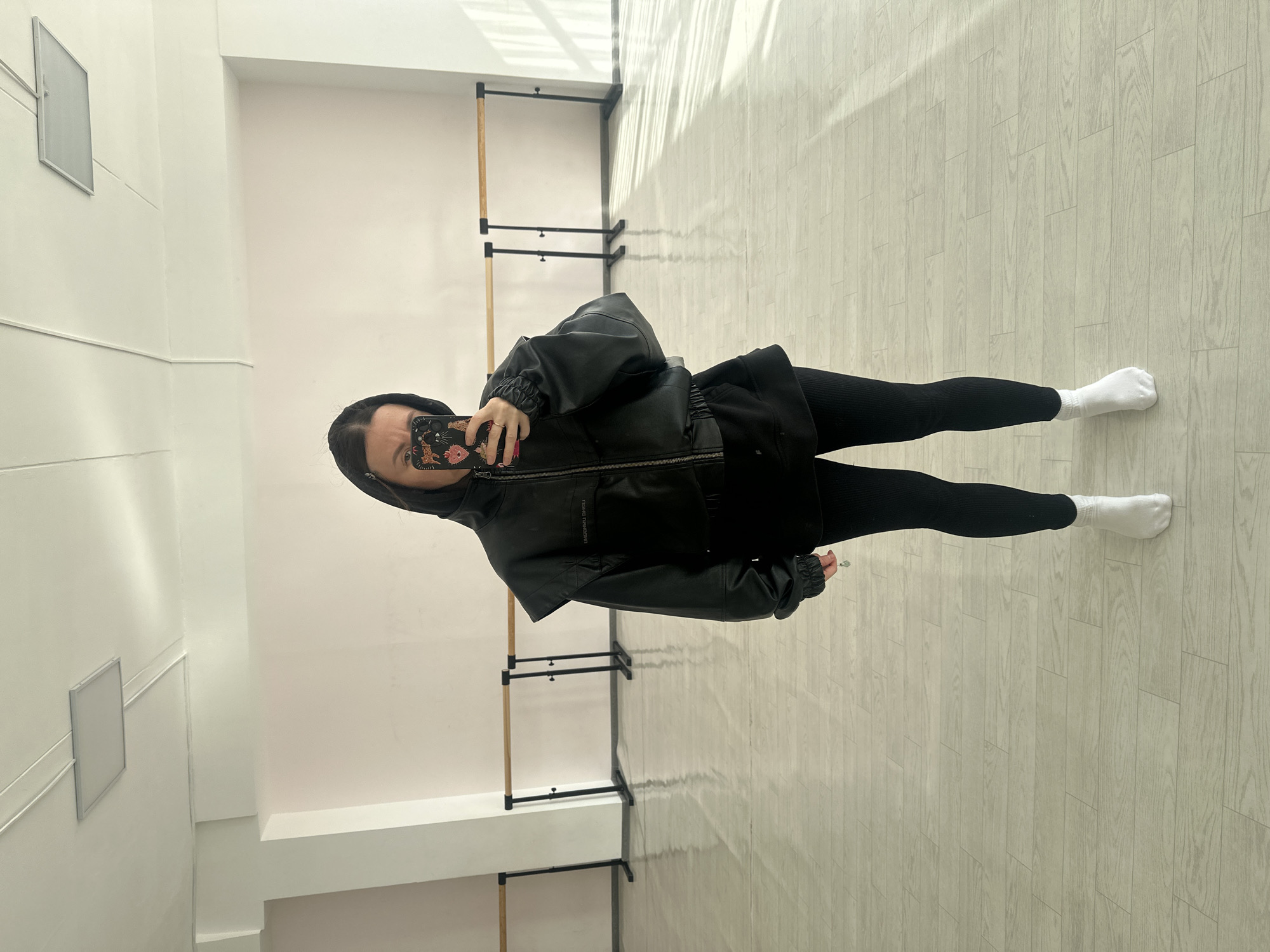 A woman stands indoors, holding a smartphone in her right hand, facing a mirror, and taking a selfie. She wears a black hooded jacket over black leggings, and white socks. The setting is a room with white walls and a light-colored wood-look floor. There are horizontal bars mounted on the walls. The location is unknown.