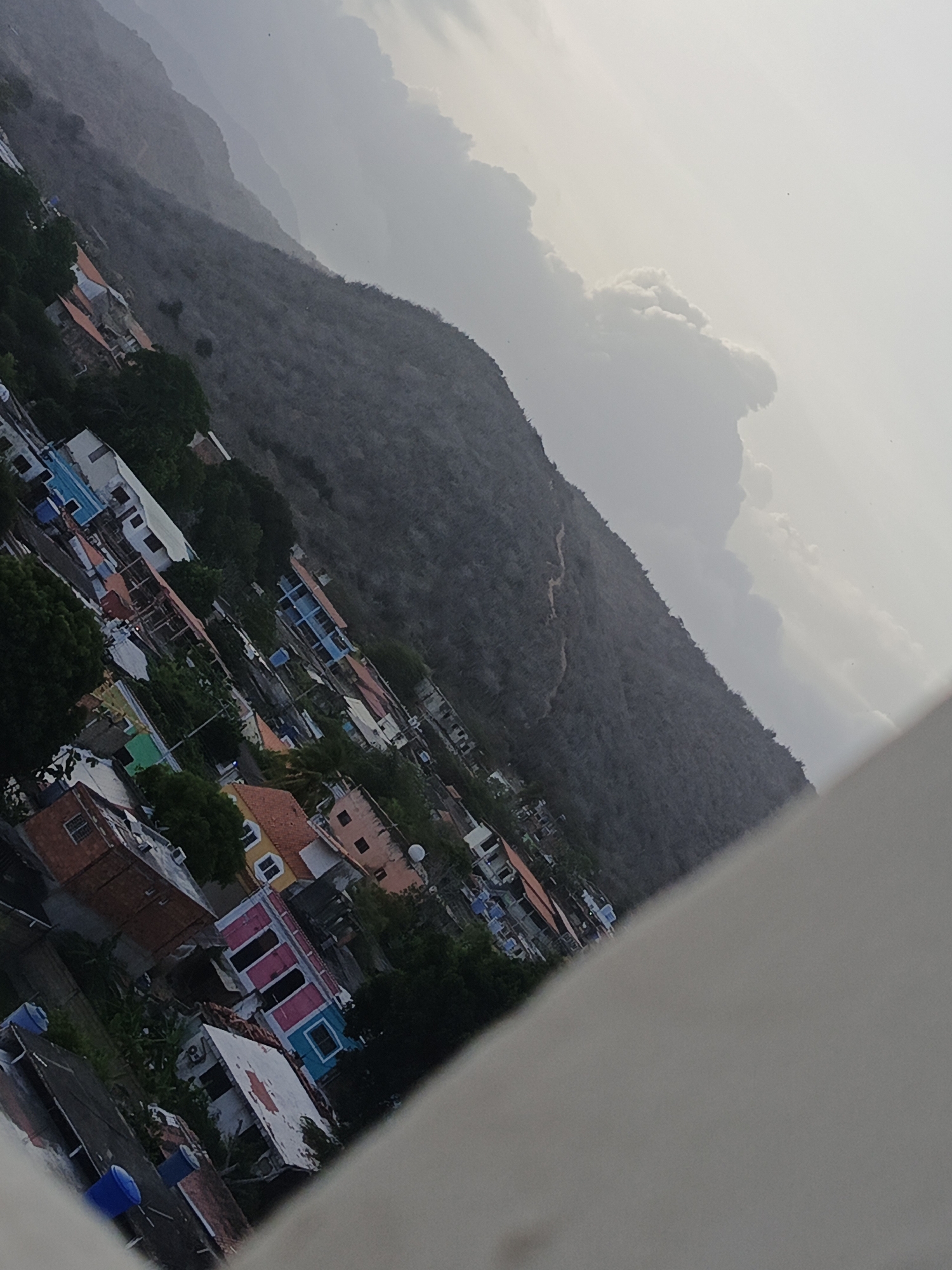 An aerial view, seemingly taken from a high vantage point, depicts the city of Los Robles in Venezuela, nestled against a mountain range partially obscured by clouds. The town consists of densely packed buildings of varying colors and sizes, interspersed with greenery. The mountainous backdrop is covered in what appears to be sparse vegetation or perhaps barren land, under an overcast sky. A portion of a white structure is visible in the foreground, potentially indicating the location from which the image was captured.