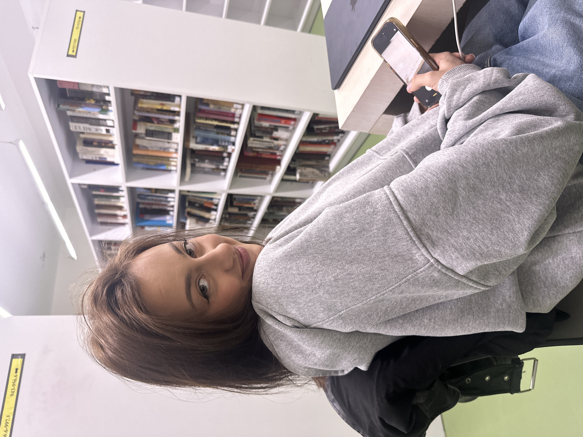 A young woman is positioned in a library. The bookshelf behind her is filled with books. She is seated at a desk with a laptop, holding a smartphone. She is wearing a gray hoodie and blue jeans. A black jacket lies on her lap. The image does not contain the information about the city of Yerevan, Armenia.