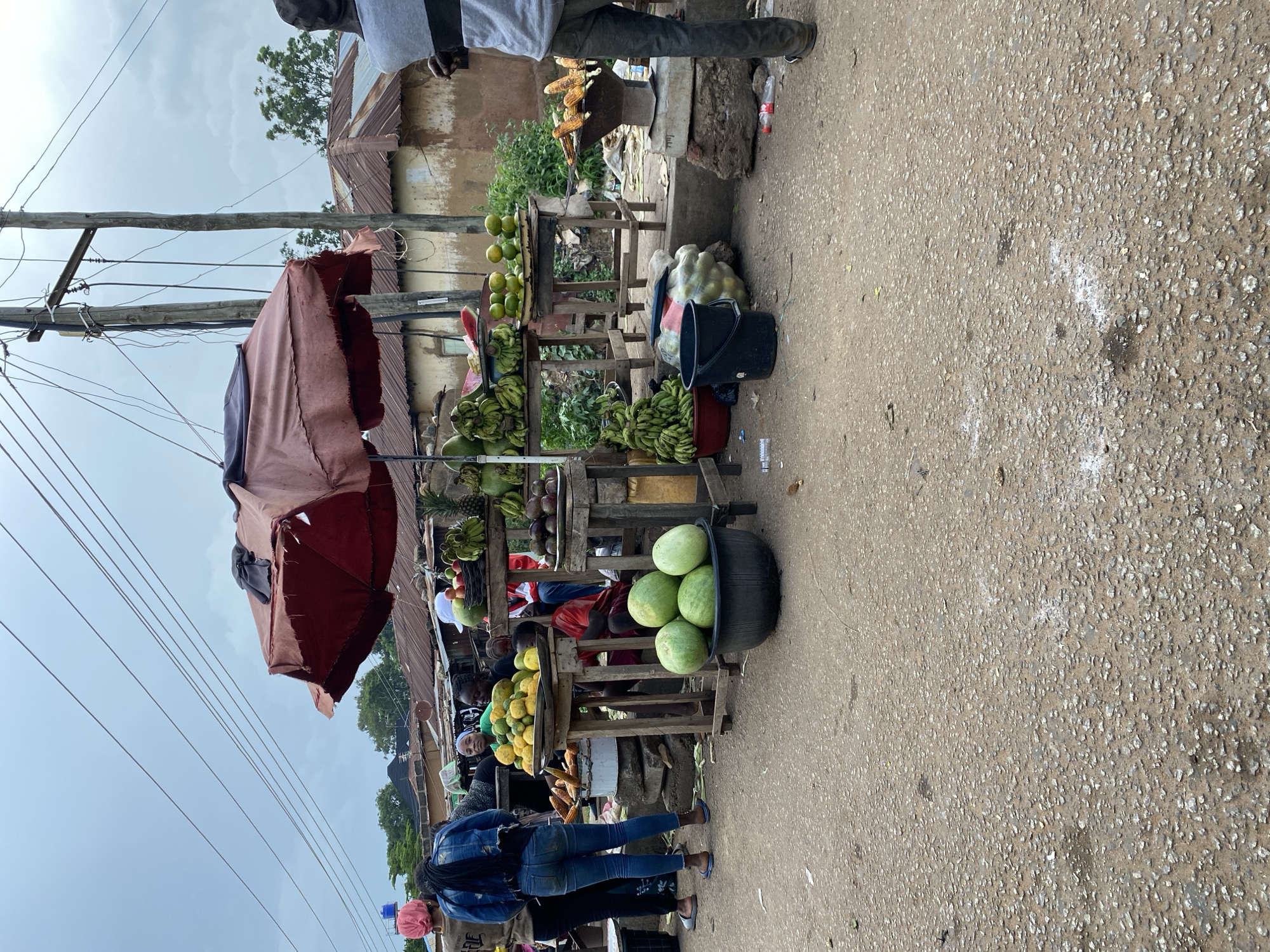 An outdoor market stall in Bukuru, Nigeria, is depicted on a cloudy day. The stall is primarily selling fruits and vegetables. Prominently featured are large watermelons in a black basin in the foreground, bunches of bananas on wooden stands, and piles of oranges and other citrus fruits. Corn on the cob is also visible, some of it appears to be grilled. Several people are present, some of whom appear to be vendors or customers. One woman in the foreground is wearing a pink headscarf and a denim jacket, and another woman next to her is wearing blue jeans. A man stands on the right side of the image, facing away from the camera. Wooden tables and stands hold the produce, and a large, faded red umbrella provides shade over part of the stall. In the background, there are buildings with corrugated metal roofs, and utility poles with power lines stretch across the sky. The ground is a rough, dirt and gravel surface.