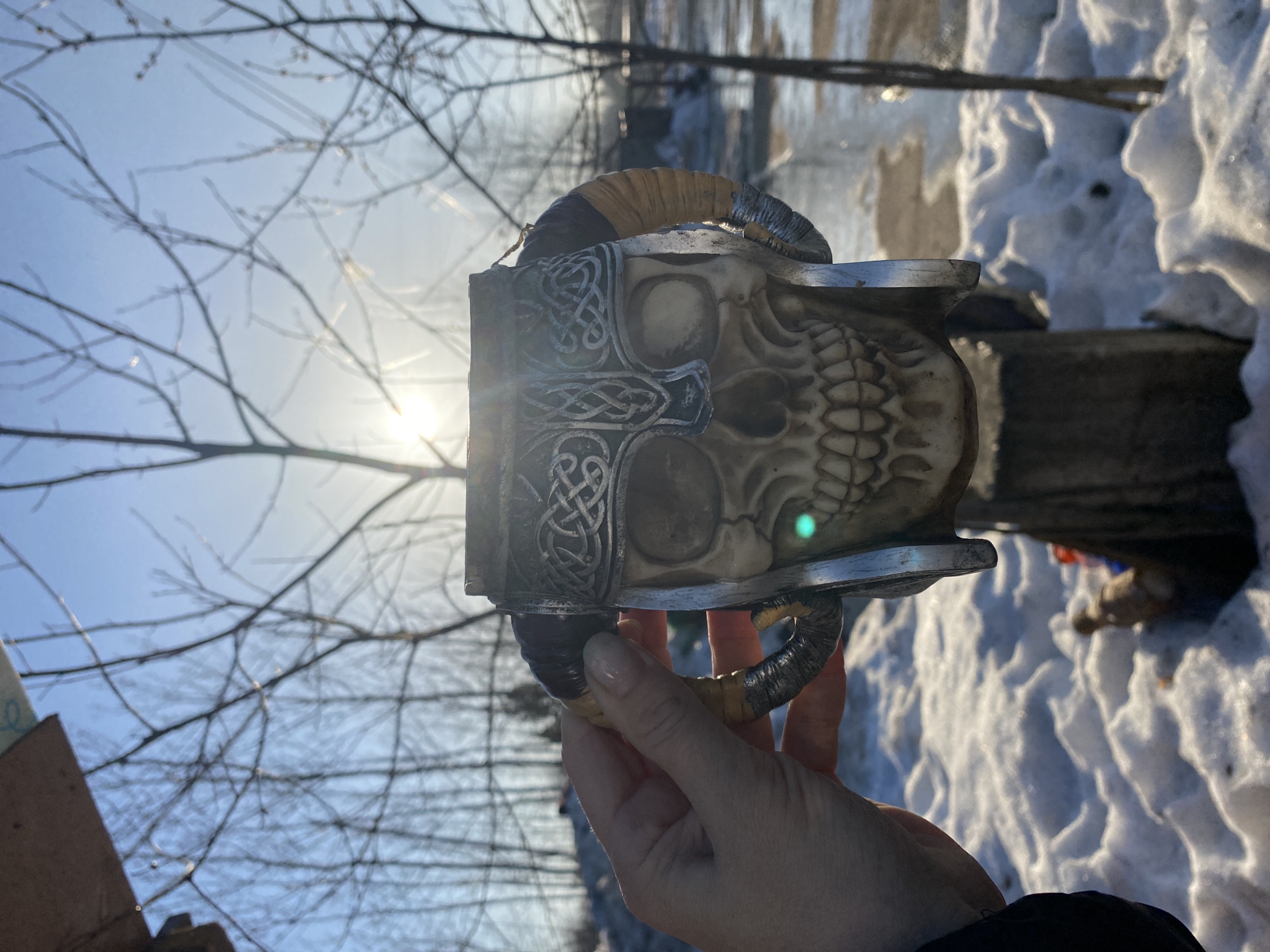 A person is holding a skull-shaped drinking vessel with horns in an outdoor setting with snow on the ground. The vessel is predominantly bone-colored with silver accents and Celtic knot designs. The environment includes bare trees against a bright sky, suggesting winter. In the background, there is water and a blurred landscape. The location is Ol'khovskoye, Russia.