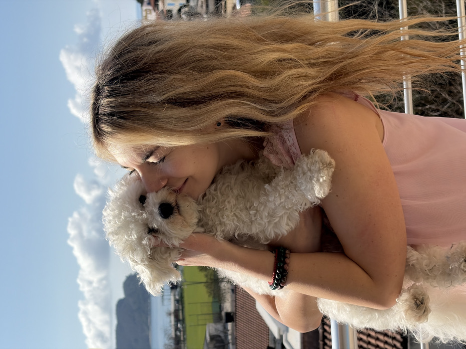 An adult female and a small, white, curly-haired dog are positioned outdoors on what appears to be a balcony, overlooking the city of Budva, Montenegro. The scene is illuminated by bright daylight under a clear blue sky with scattered white cumulus clouds.
The female, with long, wavy blonde hair, wears a light pink sleeveless top. She has several beaded bracelets on her left wrist, including black and red beads. Her face is turned towards the dog, with her right cheek pressed against its head, and her eyes closed, indicating an affectionate gesture.
The dog, a possible Bichon Frise or similar breed, is held in the female's arms, facing forward with its dark eyes and nose visible. Its front paws rest on the female's left forearm.
In the background, below a white horizontal railing, residential buildings with distinct red-tiled roofs descend towards a large body of water identified as the Adriatic Sea. A green sports field with white markings is visible in the mid-ground, bordered by fencing. A prominent, vegetated hill or mountain forms the distant coastline across the bay, consistent with the geographical features surrounding Budva.