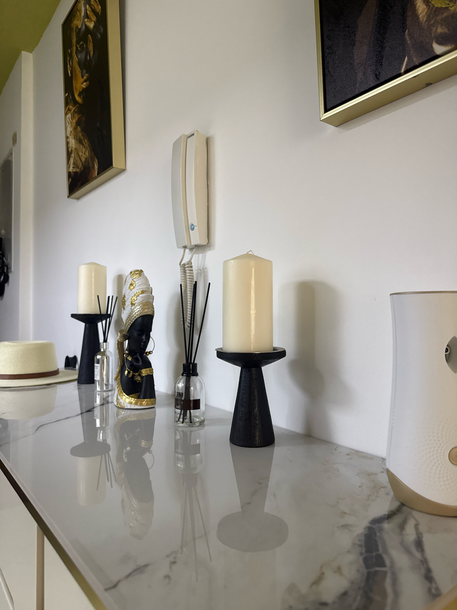 The image shows a close-up, eye-level shot of a marble-top console table in what appears to be a residential hallway in Dubai, United Arab Emirates. The console table is decorated with several items. On the left, there is a straw hat with a brown ribbon. To the right of the hat are two black candle holders, each with a white pillar candle. Between the candle holders, there is a decorative black figurine of a woman wearing a gold and white headwrap and jewelry. Several glass reed diffusers with black reeds are also placed on the table. In the background, a beige intercom system is mounted on the white wall. Further to the right, a white air freshener dispenser with a gold base is visible. The wall above the table features a framed artwork with abstract, gold and black imagery. The lighting is bright, casting soft shadows on the wall and the reflective surface of the console.