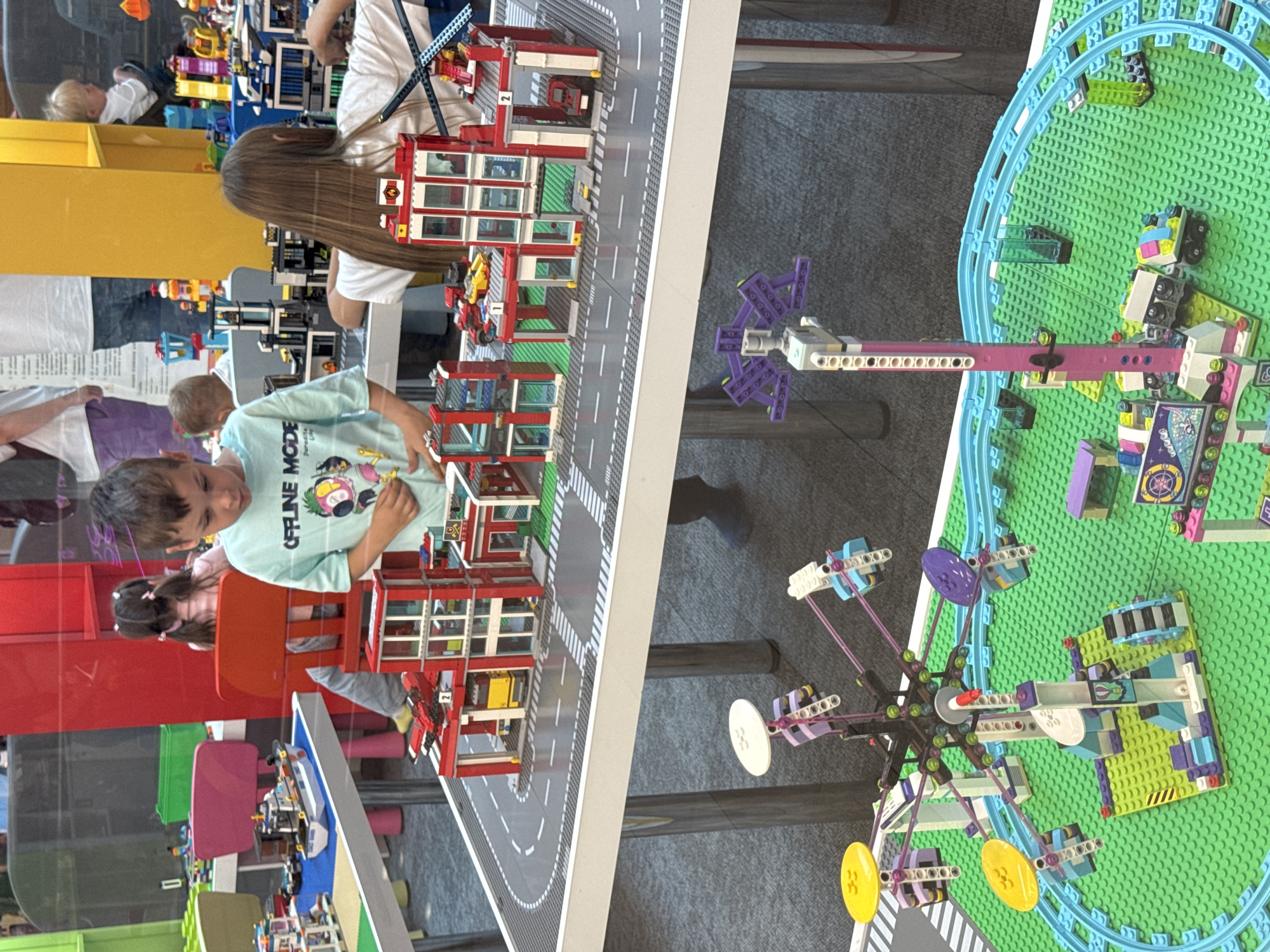 The image depicts a group of children engaged with a large Lego display, which appears to be a model of a fire station and a small amusement park. The display is set up on a white table with a grey road and green Lego baseplates. In the foreground, a boy in a light blue t-shirt with the text "OFFLINE MODE" is interacting with the Lego fire station. Behind him, a girl with long brown hair is also playing with the Lego structures. To the left, a model of a boat is visible on a blue Lego baseplate. In the background, other children and adults are present, some observing the display. The environment suggests an indoor setting, possibly a children's museum or a public exhibition space. There are red and yellow structures visible in the background, and some children are seated on chairs. The image does not contain any information that would allow for the identification of its location within the city of Ufa, Russia.