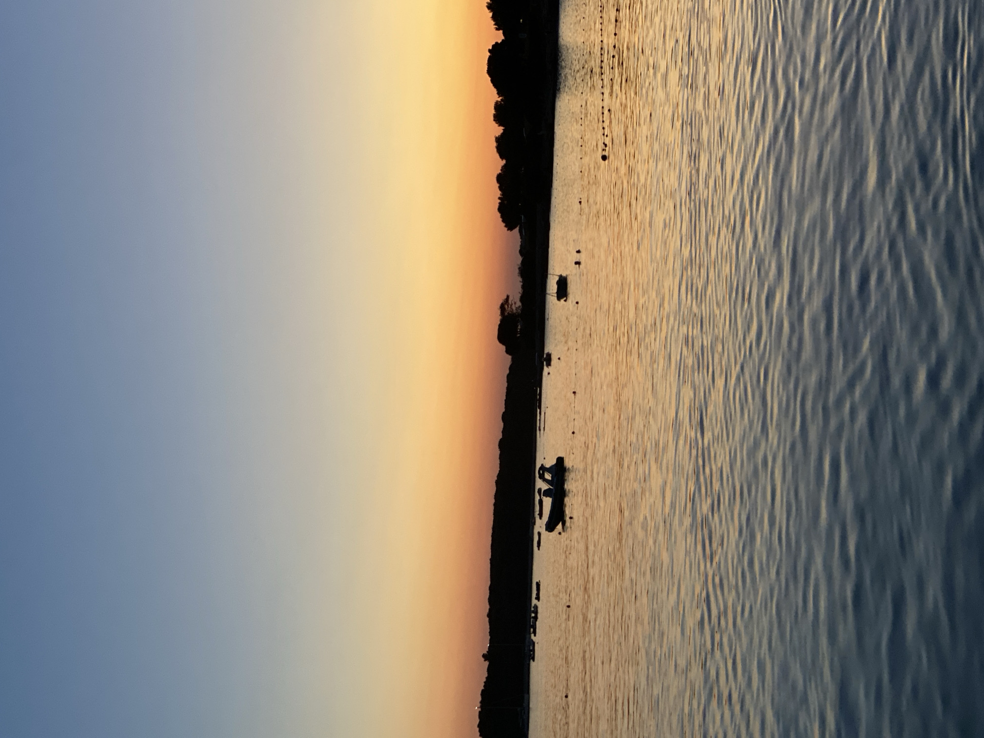 This image depicts a serene sunset over the waters near Banjole, Croatia. The sky transitions from a clear, light blue at the top to a vibrant gradient of orange and yellow near the horizon. A dark, silhouetted treeline runs along the distant shore. Several small boats are visible on the calm water, some appearing to be anchored. The surface of the water reflects the warm colors of the sunset, with gentle ripples creating a textured effect. The overall scene conveys a peaceful and tranquil atmosphere characteristic of a coastal sunset.