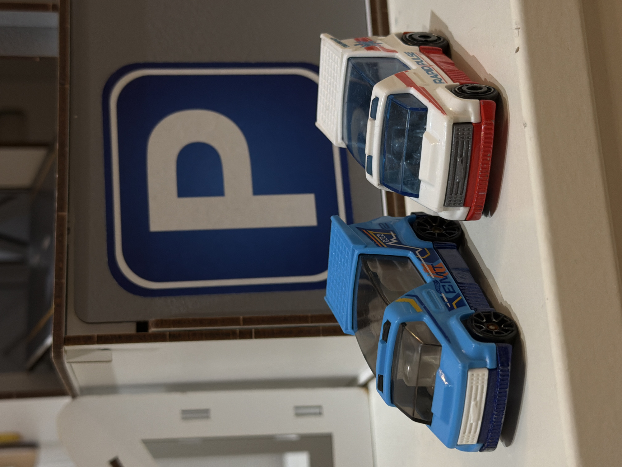 The image displays two toy cars positioned on a light-colored surface, likely a table or shelf, next to a blue and white sign. The scene is presented with a vertical orientation.
The upper toy car is predominantly white with red stripes along its sides and roof. It features the text "RAPIDPULS" printed in red on its side. This car appears to be a miniature model of an emergency vehicle, possibly an ambulance. Its front has a red bumper and a white grille, with transparent plastic windows.
The lower toy car is blue with yellow and orange accents on its sides, bearing the white text "EMT". This vehicle also resembles an emergency or utility model, perhaps a pickup truck. It has a blue bumper and a white grille, with transparent plastic windows. Both toy cars have black tires with black rims.
To the left of the cars, a prominent blue sign with a white border is visible, displaying a large white stylized letter "P", indicative of a parking sign. The area directly behind this sign is a solid gray. Further to the left, a dark brown, possibly wooden, edge runs vertically, bordering a lighter, blurry background area. The light-colored surface on which the cars rest extends towards the bottom and right of the frame, with more blurry, lighter structures visible in the distant background.
The setting appears to be indoors, with no discernible activity or event taking place, as all objects are stationary. The lighting suggests an indoor environment, possibly with artificial light. There are no people visible in the image. No specific visual cues within the image directly indicate the provided location context of Sofiivska Borshchahivka, Ukraine.