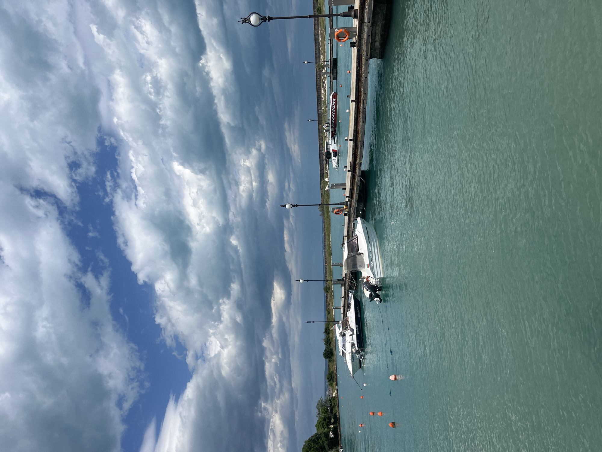 The image shows a pier with boats docked along its length, possibly in Varna, Bulgaria. The water is a light turquoise color. Several white boats are docked, secured by ropes. Orange buoys are scattered in the water. The pier has lampposts and life preservers attached. The sky is partially cloudy with patches of blue. A strip of land with greenery is visible in the background.