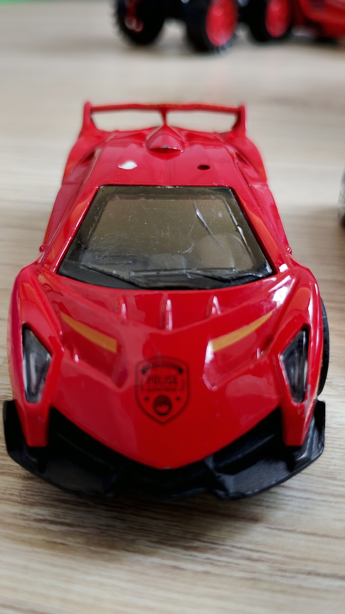 A red toy sports car is shown from a low-angle, front-facing perspective. The car is on a wooden surface and its windshield is dirty. A small circular emblem with the word "POLICE" is visible on the front of the car. The background is blurred, but shows what appears to be other toy vehicles. The setting does not provide any information that would allow for its location in Smolyan, Bulgaria.