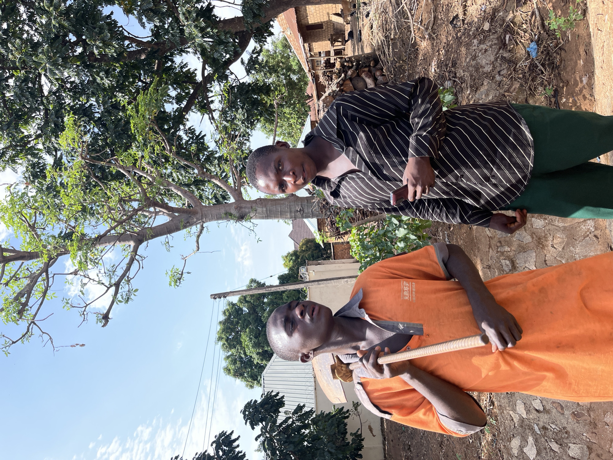 Two young men are outdoors in Barakin Momo Dogo, Nigeria. One stands in the foreground wearing an orange uniform shirt with gray trim and holding a hoe. Behind him and to the right, the second man wears a vertically striped black and white shirt and green pants. A tree looms on the left, partly obscuring the sky. Behind the two men, a dwelling with a rusty metal roof is visible amidst vegetation. A utility pole stands in the background.