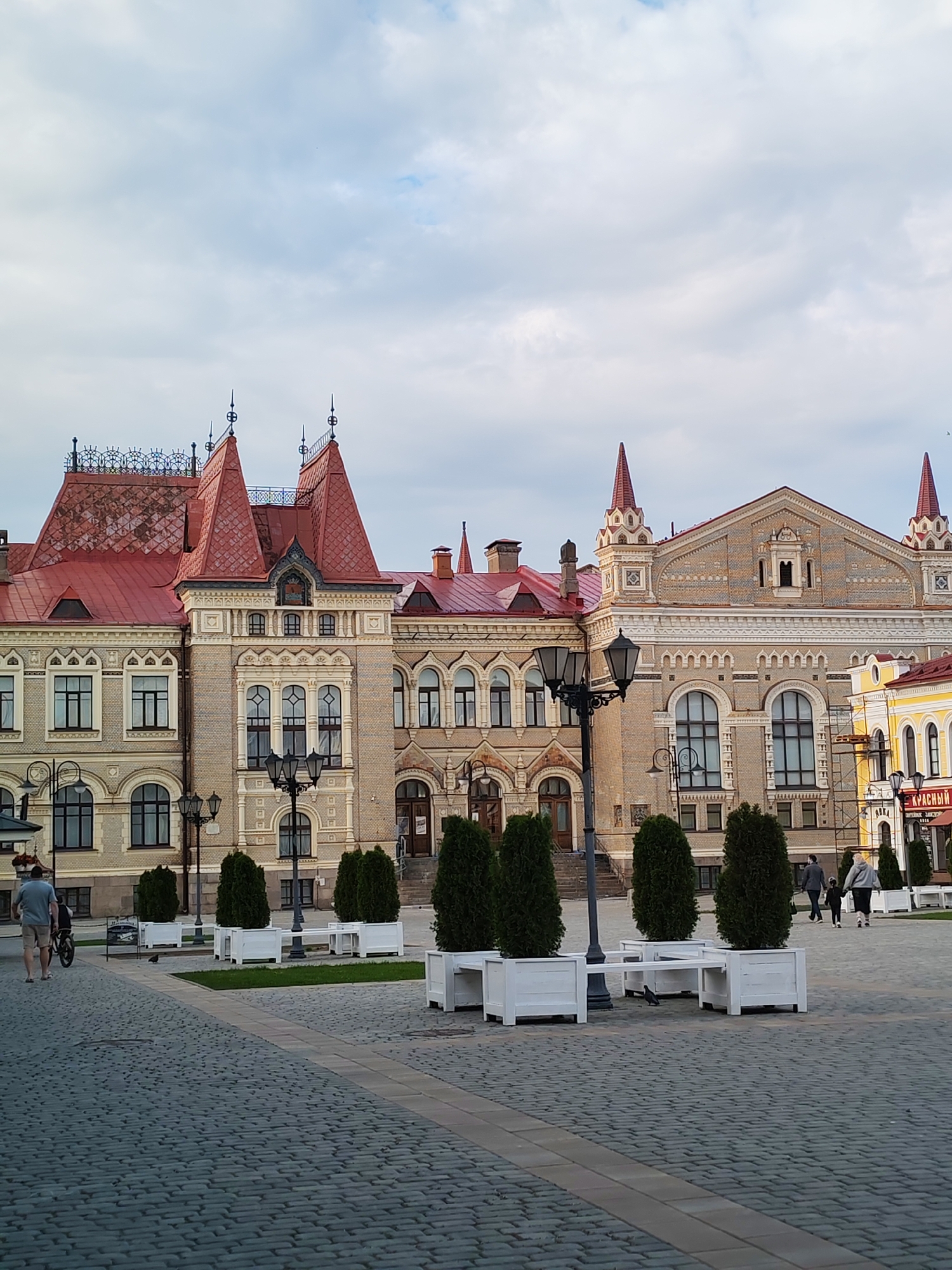 The image captures an exterior view of a grand, ornate building with a red tiled roof and intricate architectural details, likely a historical landmark, located in the city of Bol'shie Mkhi, Russia. The building features multiple towers with sharp spires, arched windows, and decorative facade elements. In the foreground, a cobblestone plaza is visible, with several modern white planters containing evergreen trees. Two individuals are walking on the plaza, one with a bicycle. The sky is overcast with scattered clouds, suggesting a temperate climate. The overall scene conveys a sense of civic pride and historical preservation in a Russian urban setting.