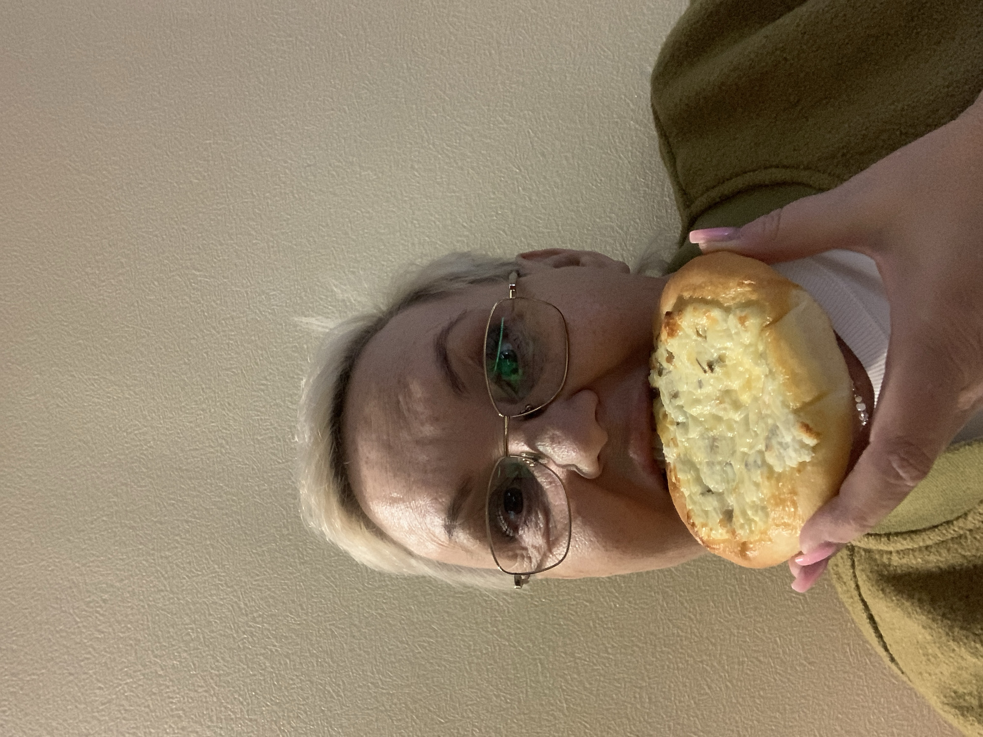 A person is holding a bread roll topped with a creamy mixture, possibly cheese or a spread. The person is wearing glasses and a yellowish-green jacket or sweater. The background is a plain, light beige wall. The location is not discernible from the image.
