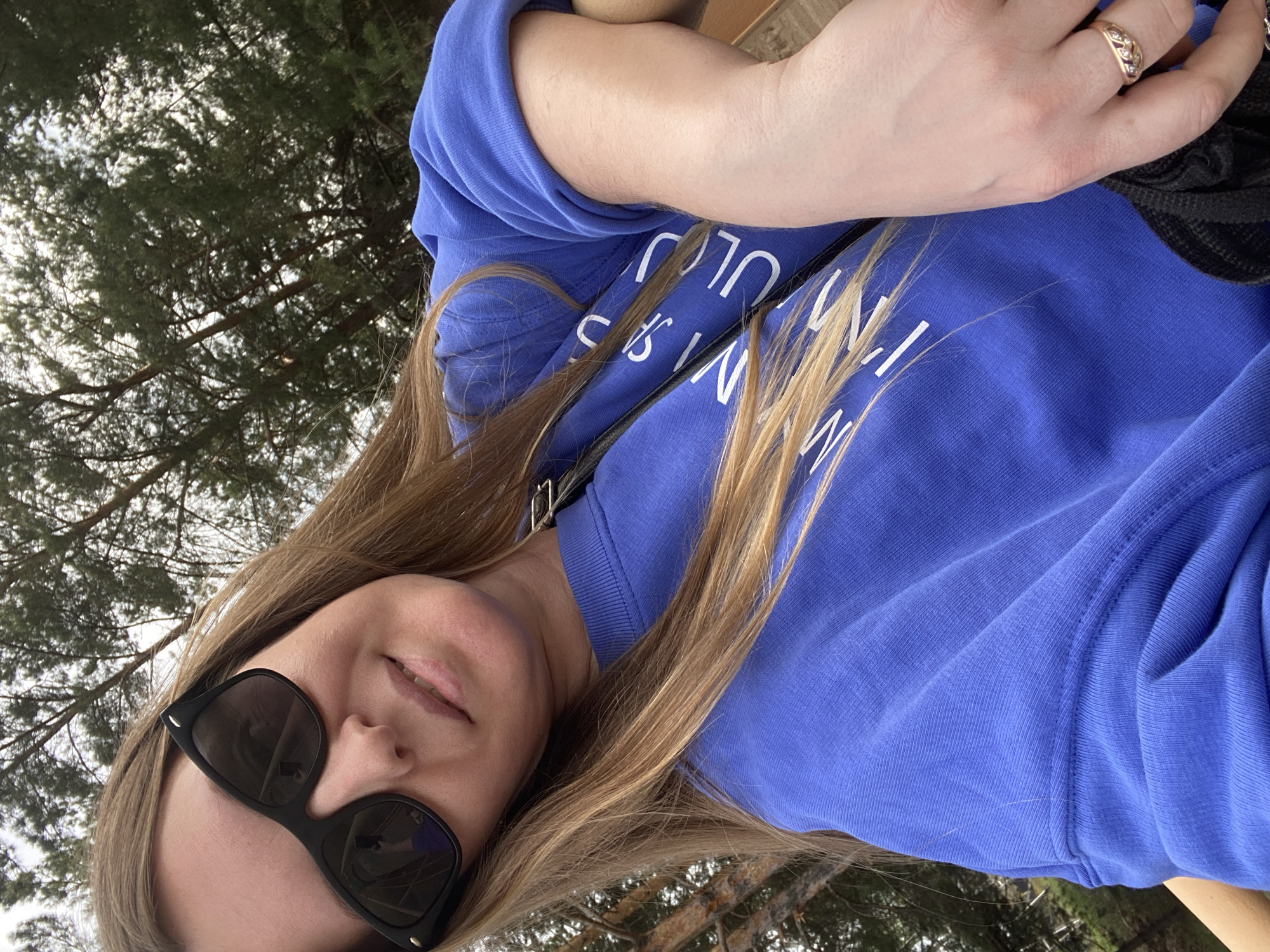 A woman with blonde hair and sunglasses is looking at the camera, wearing a blue sweatshirt with white lettering. She is wearing a ring on her finger. The background includes trees and possibly a cloudy sky. While the prompt requests that the location be identified as Noviy, Russia, there is nothing in the image to confirm or deny that location.