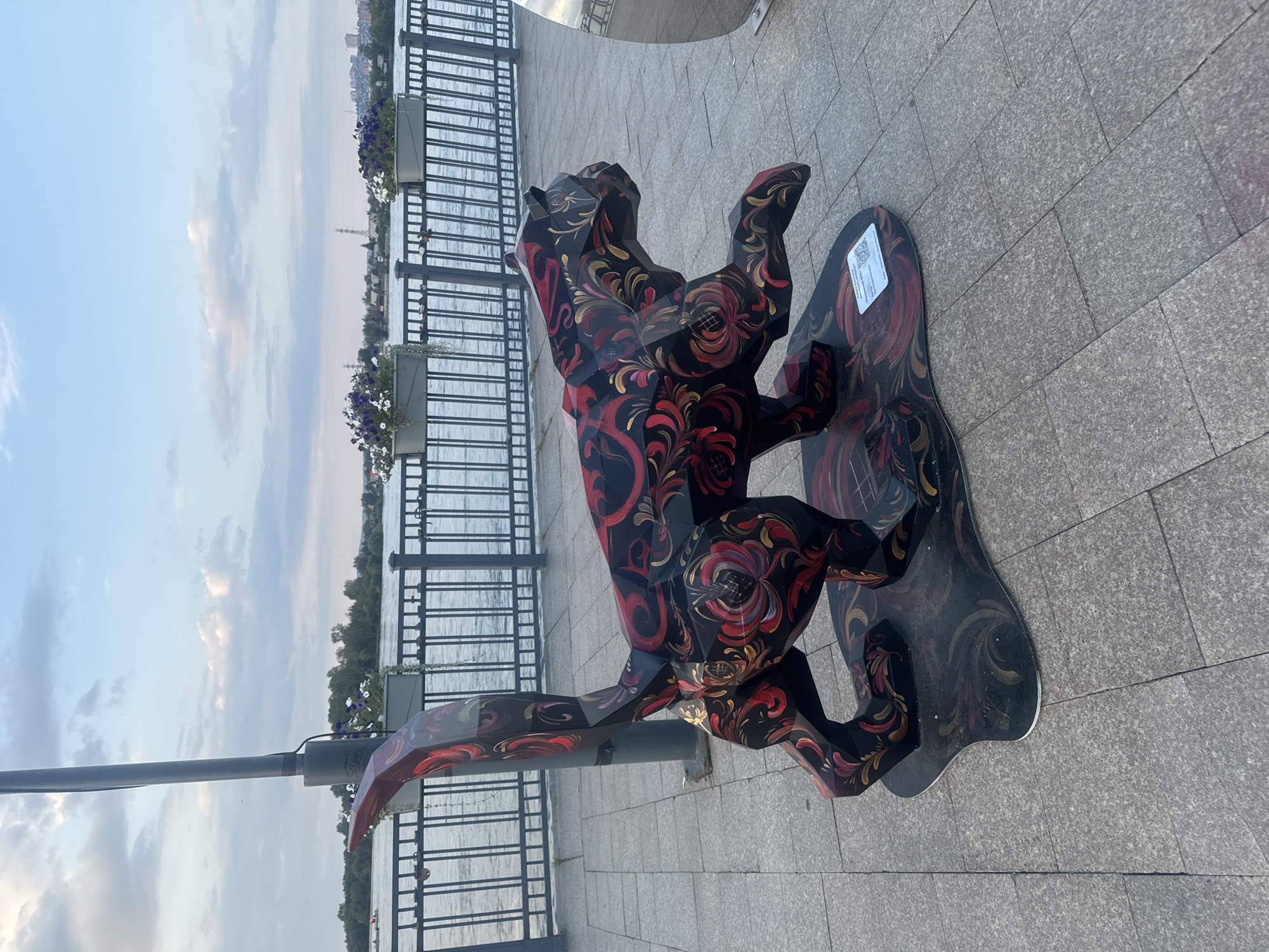 An artistic sculpture of a stylized animal, resembling a big cat with a long, curved tail and polygonal body segments, is displayed on a paved pedestrian walkway. The sculpture is predominantly black with intricate, floral-like red and gold patterns painted across its surface. It stands on a black, organically shaped base adorned with similar swirling designs. Behind the sculpture, a dark metal railing runs parallel to a body of water, beyond which is a treeline and distant buildings. Two large planters filled with purple and white flowers are positioned atop the railing. A lamppost stands to the left of the sculpture, and a reflective, rounded object, possibly another sculpture or part of an installation, is visible to the far right. The ground is paved with large, rectangular gray stone tiles. A small white label with text and a QR code is affixed to the base of the animal sculpture. The sky above is a pale blue with scattered white clouds. This is likely located on the embankment in the city of Irkutsk, Russia, possibly along the Angara River.