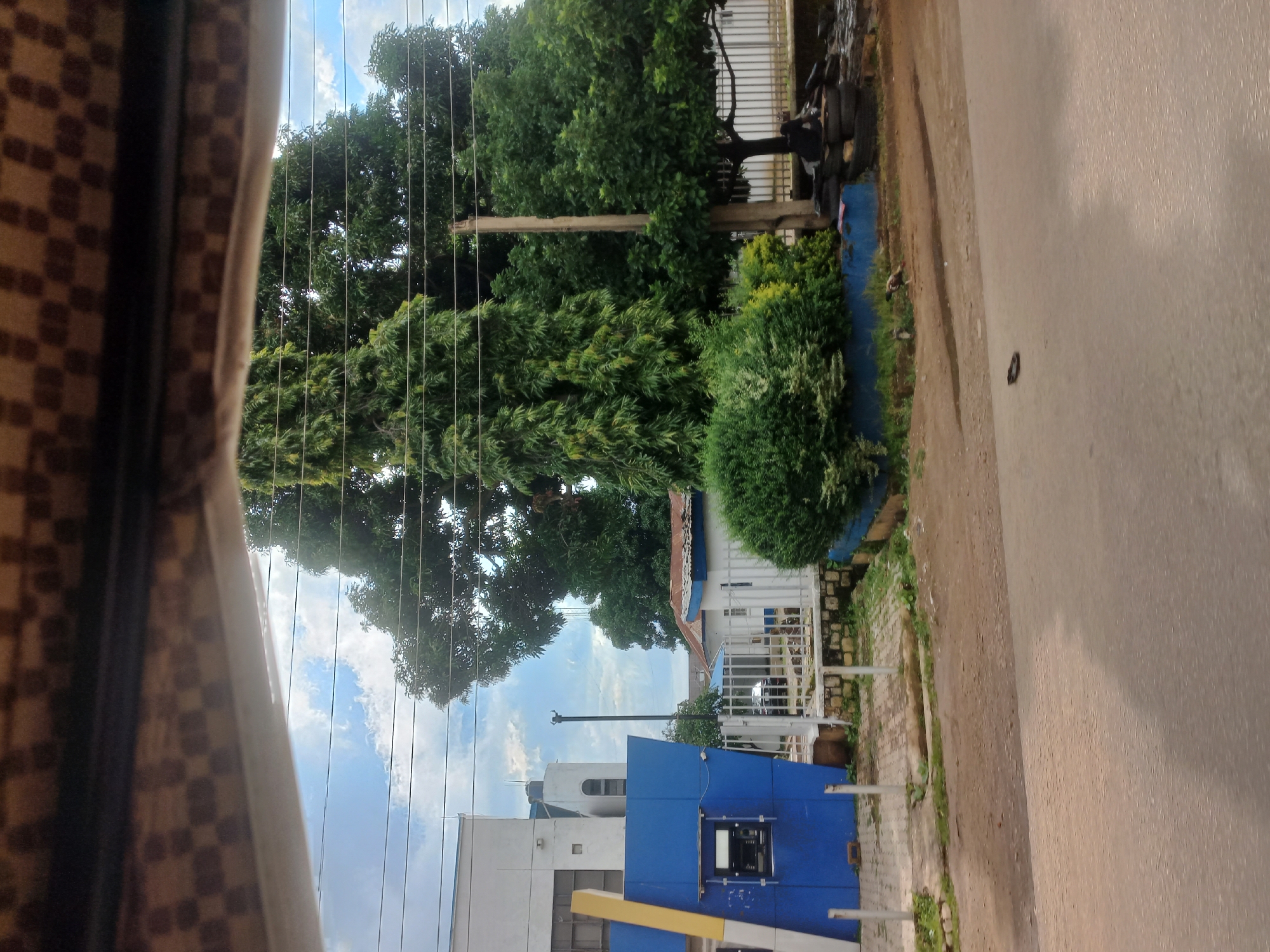The image depicts a street view in Jos, Nigeria. In the foreground, a paved road is visible. Behind the road, a dirt path leads to a blue building with a yellow vertical accent, likely a bank or an ATM kiosk. Several power lines stretch across the sky, intersecting with tall trees. Adjacent to the blue building, a white metal fence encloses a courtyard with a few small buildings and overgrown vegetation. A concrete pole stands near the fence, and several tires are stacked at its base. The sky is partly cloudy. The scene appears to be captured from a moving vehicle, as suggested by the blurred foreground and the framing of the image.