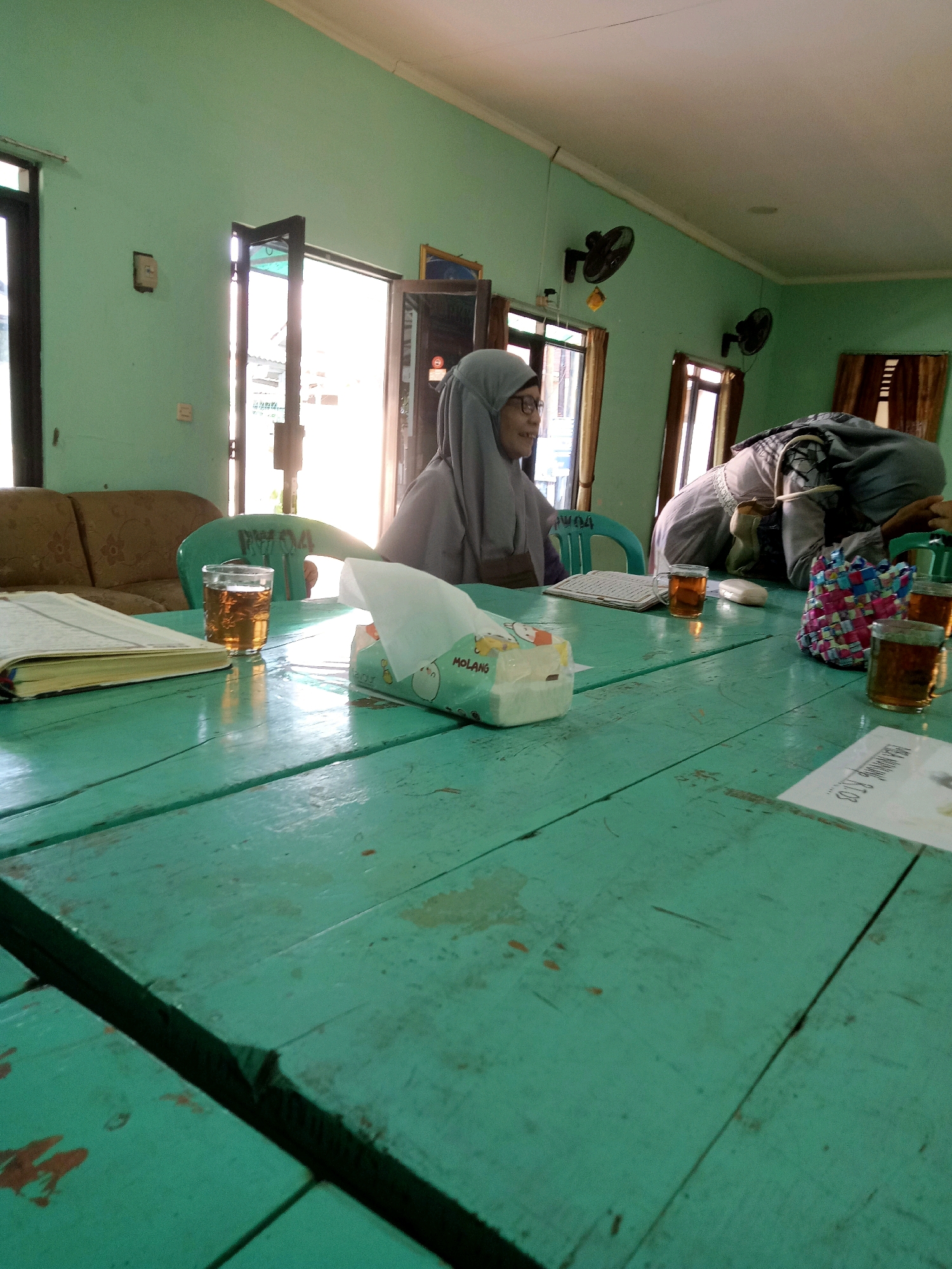 The image depicts an indoor scene at what appears to be a simple room, possibly a community space or a dwelling, in Cirebon, Indonesia. A long, weathered, turquoise wooden table occupies the foreground, with several items scattered across its surface. To the left, an open book, likely religious in nature given the context and attire of the people, rests near a glass of amber liquid, presumably tea. A tissue box with the brand "Pang" is also visible on the table.
Two individuals are present. One, wearing a light grey hijab and glasses, is seated on the left side of the table, facing away from the camera and towards the right. The second individual, also in a hijab and wearing a grey garment with a patterned overlay, is bent over the table on the right side, seemingly engaged with something on the surface. Their faces are not clearly visible.
The room's walls are painted a light mint green. Two wall-mounted fans are visible on the wall in the background. A sofa is partially seen behind the seated individual. Natural light streams in from windows or doorways at the far end of the room, creating bright areas and casting shadows. The overall impression is a functional, unpretentious environment.