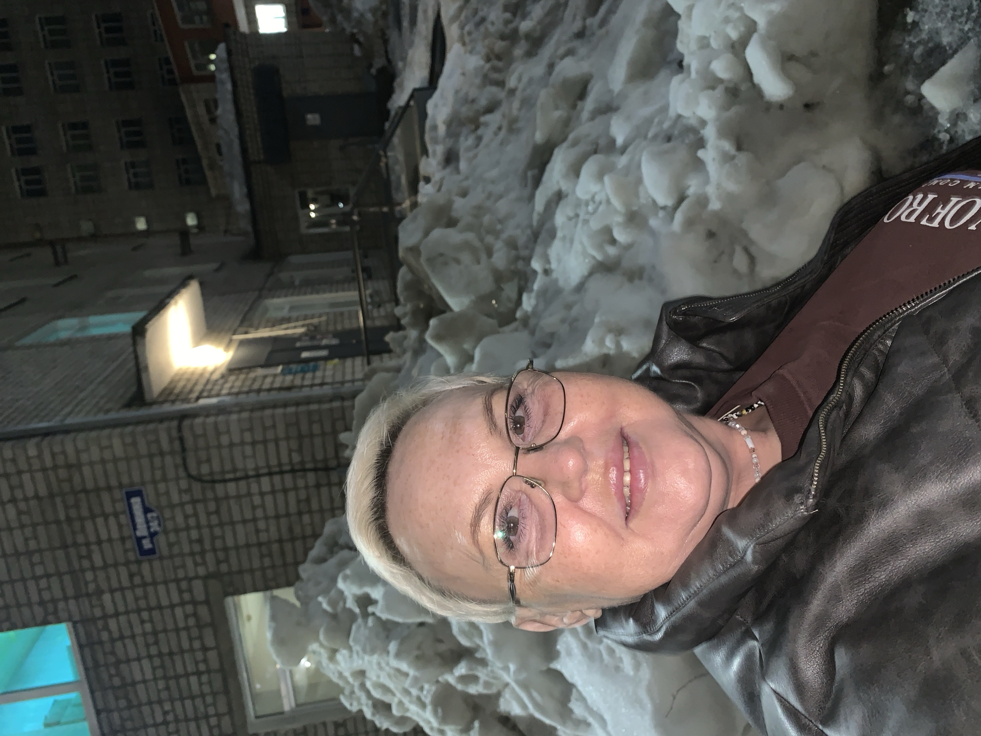 The image displays a close-up, waist-up portrait of a female individual taken outdoors at night on Lenin Street (ул. Ленина), address 24/2, in Chaikovsky, Russia. The subject is a middle-aged woman with short, light blonde hair and metal-rimmed glasses, smiling directly at the camera. She is wearing a dark brown leather-like jacket over a reddish-brown top, with a light-colored beaded necklace visible around her neck.
The background is dominated by extensive, irregularly shaped piles of snow covering the ground. To the left of the subject, a multi-story brick building is visible, featuring a blue street sign with white text "ул. Ленина 24/2". A window on this building emits a turquoise light, and a yellow-orange light fixture illuminates an overhang further back. In the distant right background, another dark, multi-story building is faintly visible against the night sky, with several windows emitting light. The scene is illuminated by artificial light sources.