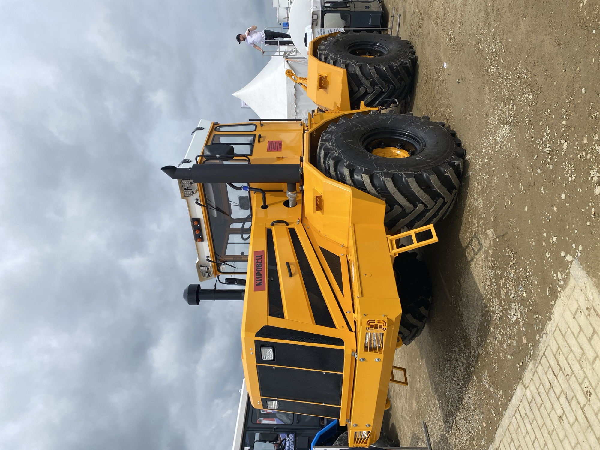 The image shows a large, yellow Kirovets tractor parked on a dirt and gravel surface with a brick path on one side. The sky is overcast. In the background, there is a white tent and a person standing on a raised platform. The tractor is the main subject of the image, with its large tires and prominent exhaust pipe. The location is potentially in Voronezhskaya, Russia, based on the tractor's brand, although there is no other evidence in the image to confirm it.