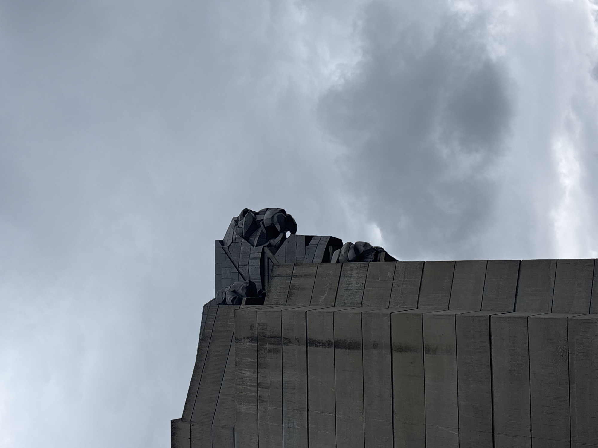 The image displays a close-up of the upper section of a large, dark, brutalist-style building or monument against an overcast sky. The structure is characterized by its monumental scale, angular design, and textured surface, likely made of concrete or dark stone.
Prominently featured at the top-left edge of the structure is a detailed, stylized sculpture of what appears to be a powerful bird, possibly an eagle or a similar bird of prey. This sculpture exhibits sharp, geometric lines, forming its head, beak, and body, with visible claws gripping the architectural elements below it. Further along the top edge to the right, other abstract or stylized sculptural forms are integrated into the architecture, contributing to the edifice's imposing character.
The sky is filled with a mixture of light and dark grey clouds, indicating an overcast day with diffuse lighting. There are no human figures or activities visible in the frame. The overall impression is one of solemnity, power, and architectural grandeur. No discernible text is visible anywhere in the image.