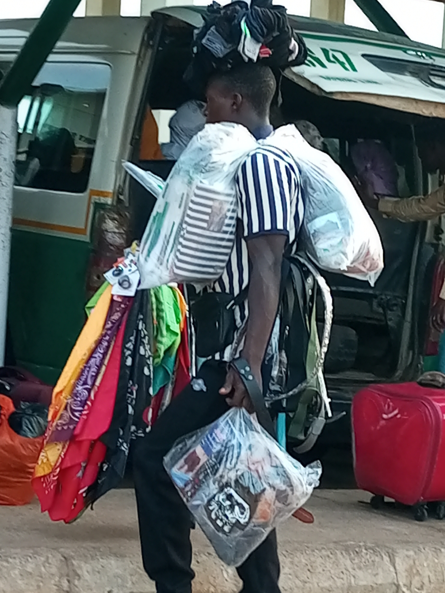 A Black male, dressed in a blue and white striped shirt and black pants, is seen from the waist up, standing in what appears to be a bus park in Gombe, Nigeria. He is carrying an assortment of goods, including two large plastic-wrapped bundles of items, one on his back and another in his hand. He also has a stack of clothes precariously balanced on his head, and a collection of colorful fabrics hanging from his right side. In the background, a white bus with green trim is visible, with its door open and other people around. To the right, a bright red suitcase is on the ground, suggesting a travel or transport scene. The ground beneath is a rough, concrete surface.