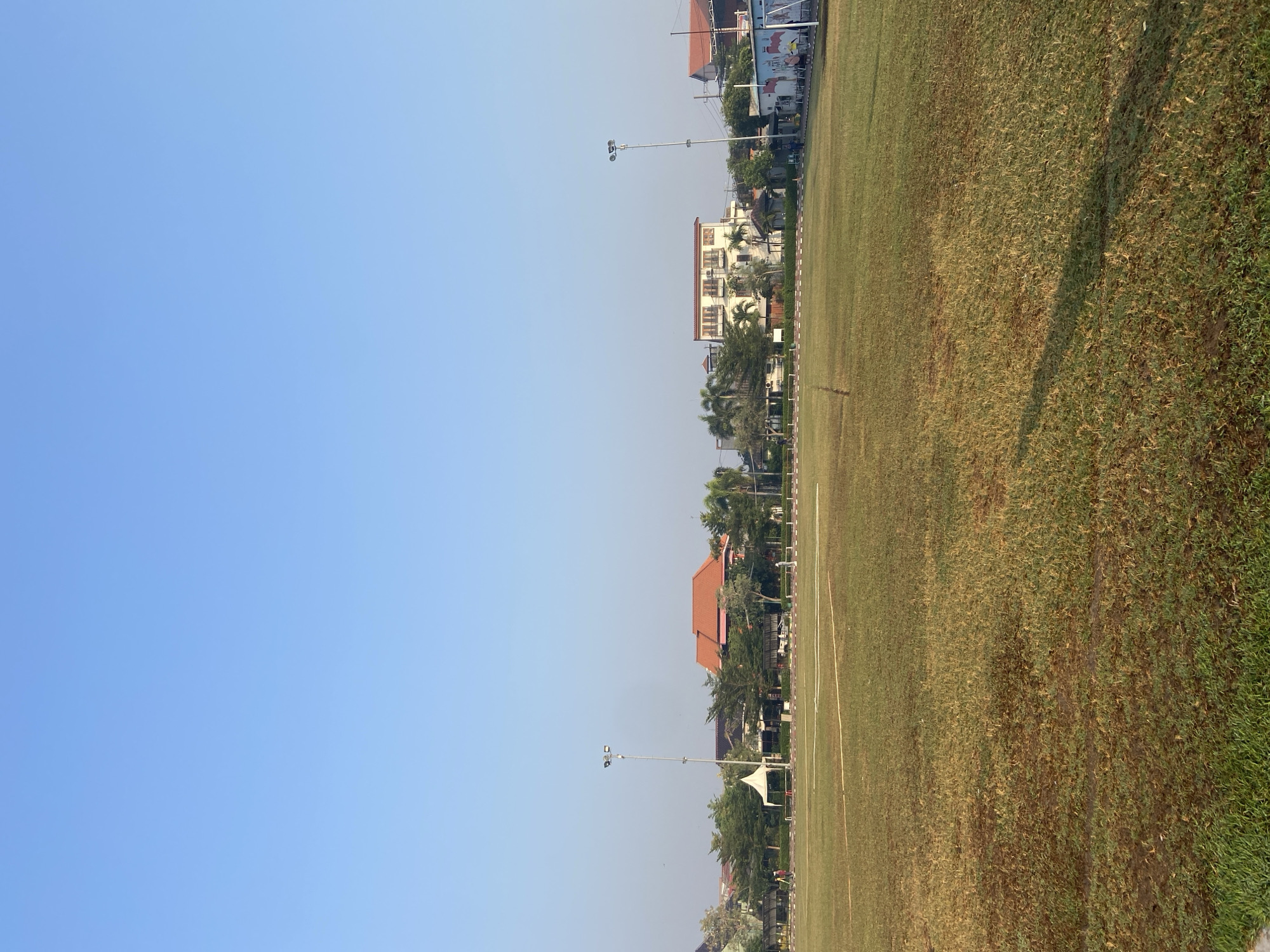 An outdoor, full-shot image depicts a grassy field with buildings and trees in the background under a clear blue sky. The field appears to be a sports field, with white lines marking boundaries. In the background, several structures are visible, including houses with red-tiled roofs, a multi-story building with balconies, and smaller utility buildings. Trees and palm trees are scattered throughout the background, providing greenery. Two tall lampposts are positioned on either side of the field, suggesting it may be used for evening activities. A few people are visible in the distance, walking or standing near the buildings. The overall impression is a calm, daytime scene in a semi-urban or suburban area. The image does not provide any specific clues about the location in Sumorame, Indonesia, beyond the general context of a field and buildings.