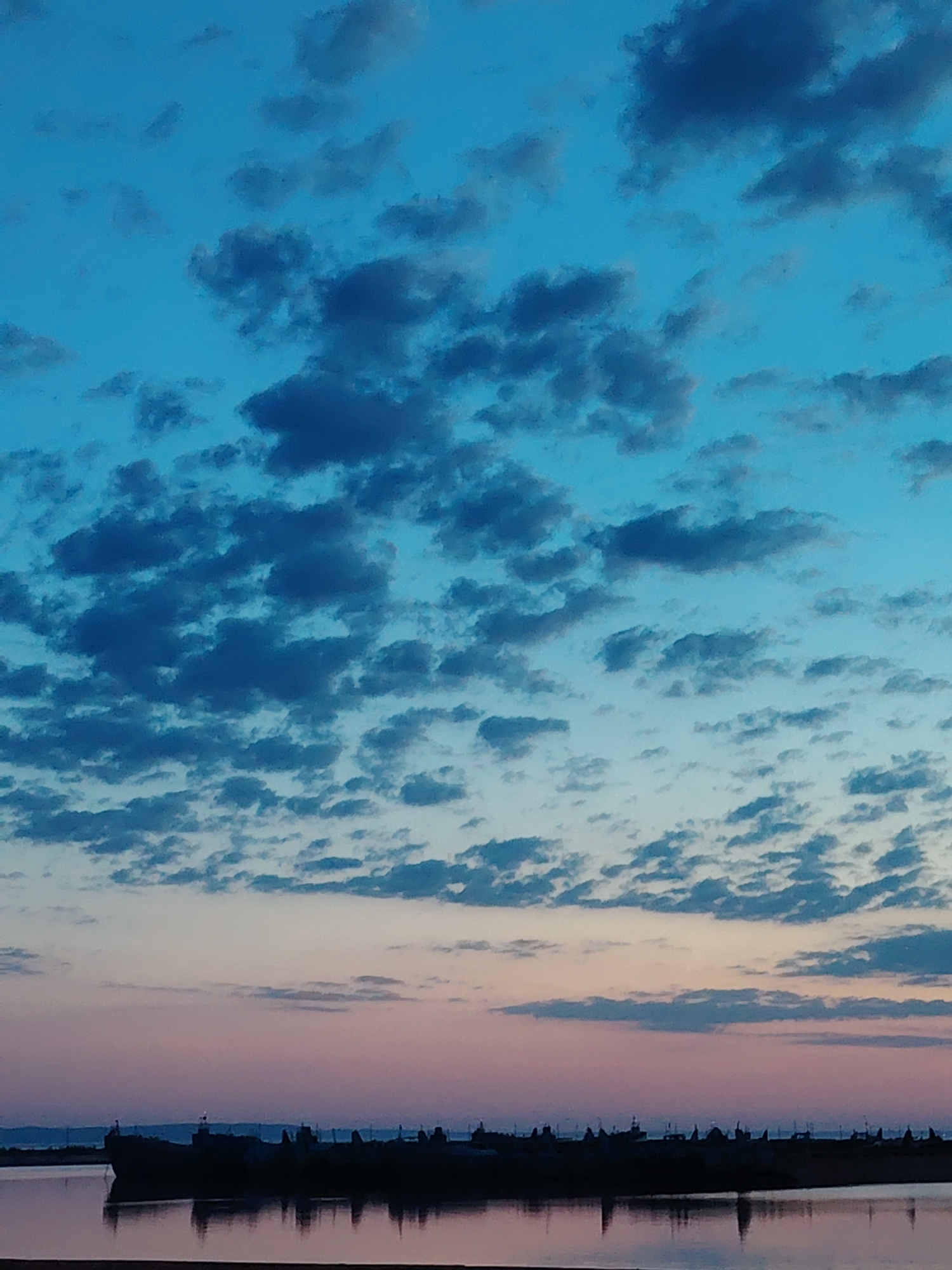 The image captures a twilight scene in Chaikovsky, Russia, near a body of water. The sky is predominantly blue, transitioning to a pinkish-purple hue towards the horizon. Numerous small, dark clouds are scattered across the sky. The silhouette of what appears to be a long structure with multiple pointed elements is visible along the horizon. The water reflects the silhouettes and the colors of the sky, creating a mirror-like effect.