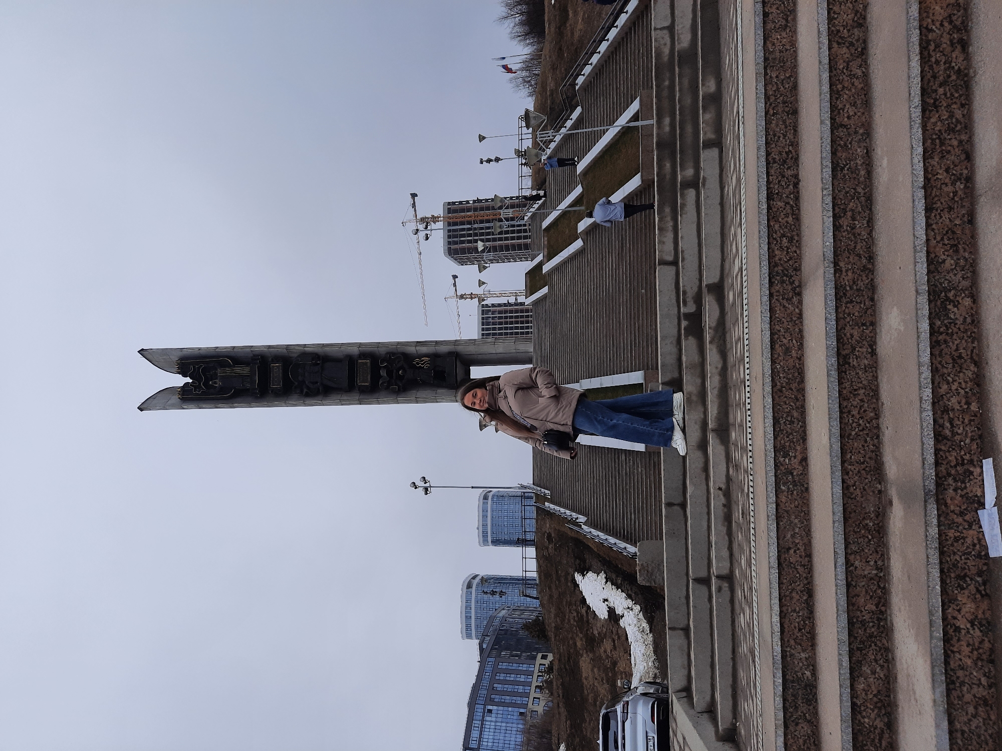 The image shows a woman standing on a staircase in Izhevsk, Russia, with the "Forever with Russia" monument in the background. The staircase has alternating concrete and brown granite steps. The woman is wearing a light brown jacket, jeans, and white sneakers. She is holding a black purse. In the background, there are buildings under construction with cranes, as well as other buildings and patches of snow on the ground. The sky is overcast.