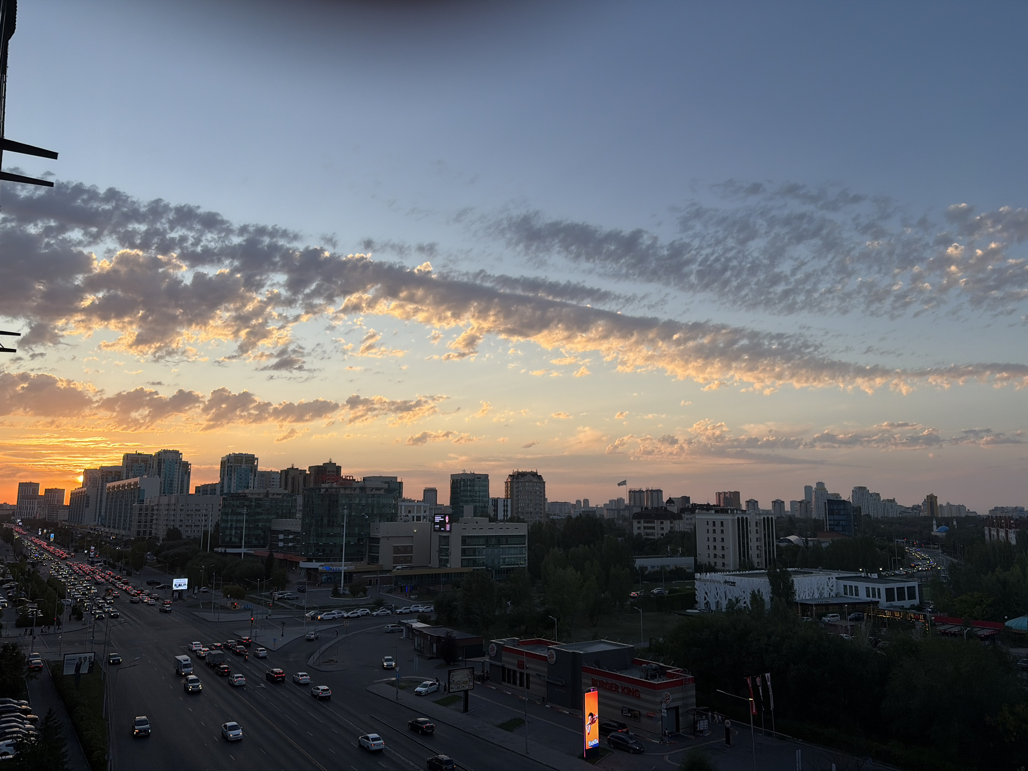The image depicts a wide-angle view of a cityscape at sunset in Astana, Kazakhstan. The sky is a gradient of light blue, transitioning to warm orange and pink hues near the horizon where the sun is setting behind a cluster of tall, modern buildings. Wispy, elongated clouds are illuminated by the setting sun, casting a warm glow across the sky.
In the foreground and midground, a multi-lane road is congested with traffic, indicating a rush hour or significant vehicle flow. Numerous cars and trucks are visible, moving in both directions. Adjacent to the road are sidewalks and various buildings, including commercial establishments like a Burger King with its distinctive signage. Green trees line some sections of the road and surrounding areas, providing a natural contrast to the urban environment.
The skyline is dominated by contemporary high-rise buildings, varying in architectural style and color. Some buildings feature glass facades that reflect the sky and surrounding lights. The overall impression is of a bustling, developing city under a dramatic evening sky. The perspective suggests the photo was taken from an elevated vantage point, possibly a tall building or balcony, overlooking the city.