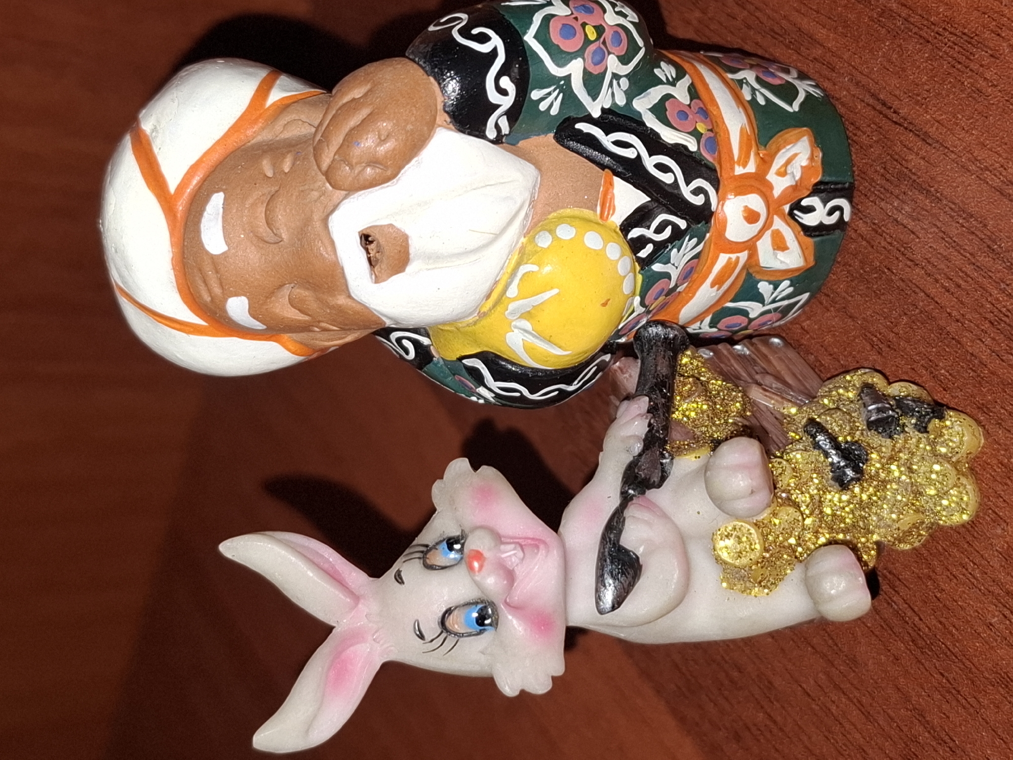 The image displays two figurines on a wooden surface. On the left is a white bunny figurine with pink ears, blue eyes, and a wide smile, holding a silver spoon. The bunny is positioned next to a cluster of gold glitter and black elements that resemble coins or nuggets. To the right stands a figurine of an elderly man wearing a white and orange turban and a green robe with intricate floral patterns in pink, white, and black. The man has brown skin, a white beard, and his hand is near his mouth. The figurines appear to be decorative items. The scene is captured in a close-up, indoor shot, with a shallow depth of field that blurs the background. There is no indication of the city of Almaty, Kazakhstan, or any specific location within it.