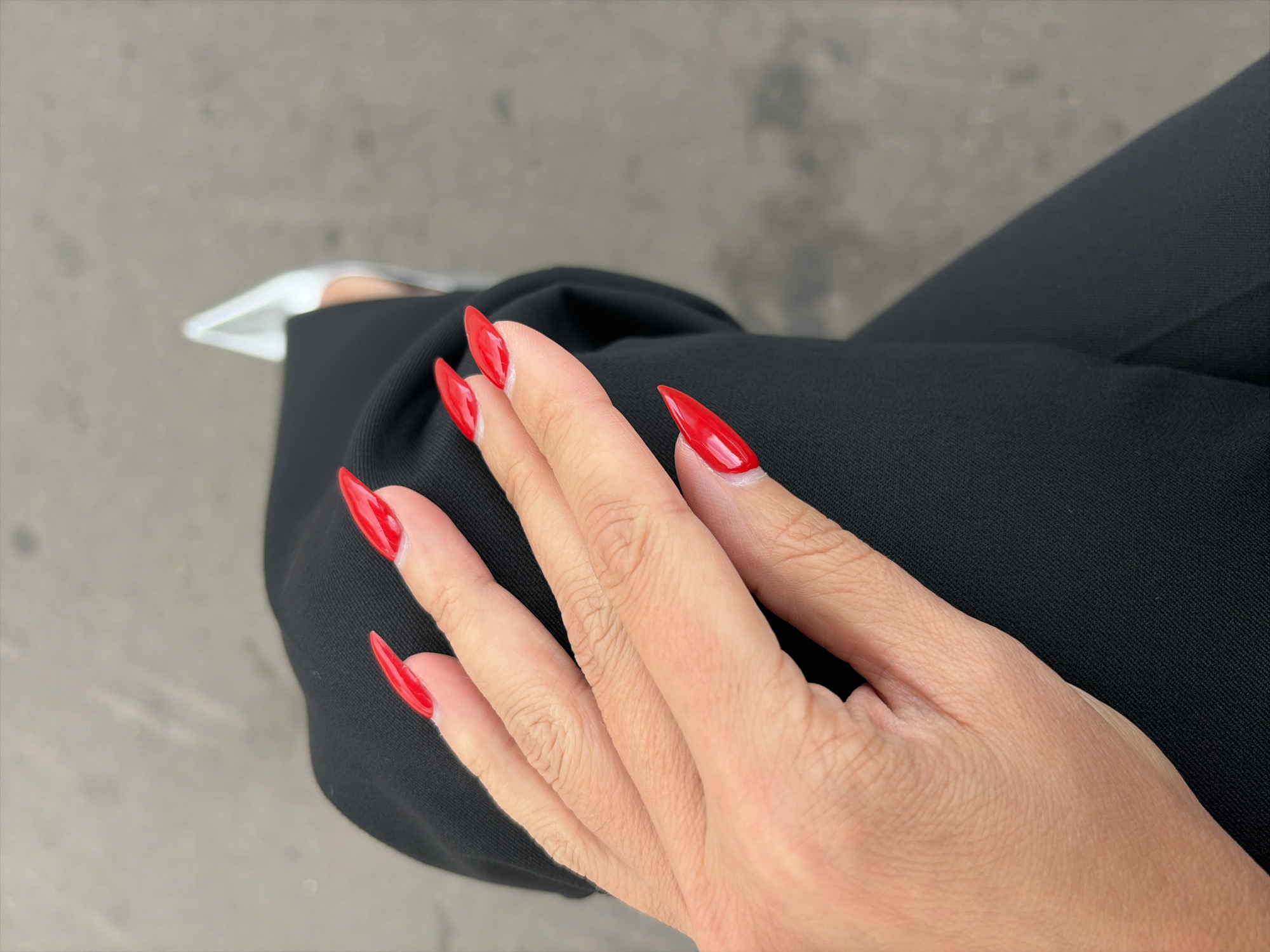 A close-up, high-angle, outdoor shot shows a person's left hand resting on their lap, which is covered by black fabric. The hand has long, pointed fingernails painted a glossy, vibrant red. The fingernails are filed into a stiletto shape. The person's fingers are slightly curled. In the upper right corner, the toe of a white, pointed shoe is visible. The background is a textured, gray surface, possibly pavement or a sidewalk. The lighting suggests it is daytime.