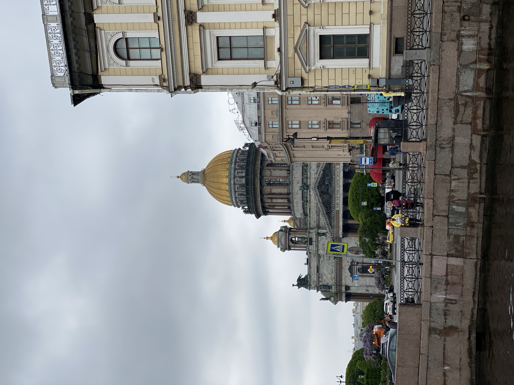 The image captures Saint Isaac's Cathedral in Saint Petersburg, Russia, under a cloudy sky. The golden dome of the cathedral dominates the scene. Adjacent buildings with ornate facades are visible. A stone embankment runs along the edge of a waterway. People walk along the embankment near a black metal fence. Cars are parked nearby. Street signs and foliage are present.