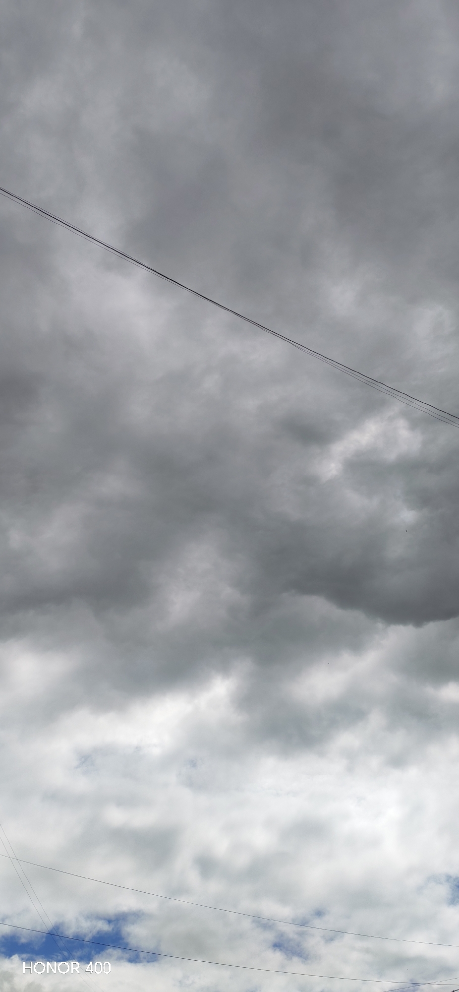 The image depicts a cloudy sky with varying shades of gray, ranging from dark, heavy clouds overhead to lighter, whiter clouds near the bottom of the frame. A thin, dark wire runs diagonally across the upper portion of the image. In the lower left corner, "HONOR 400" is visible, indicating the device used to capture the image. Patches of blue sky are visible amidst the clouds in the bottom section. The location is Noviy, Russia.