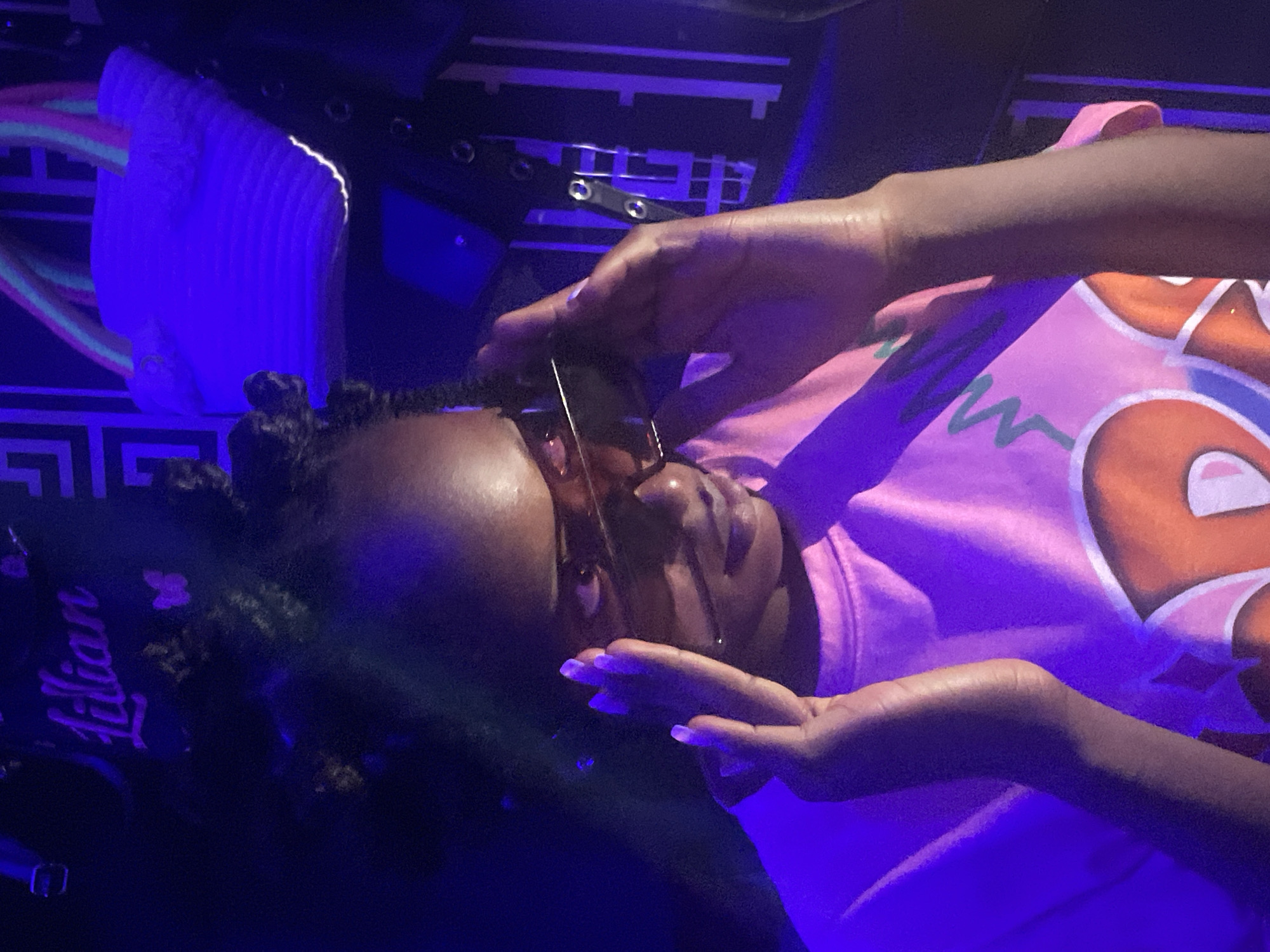 A person with dark skin and dark hair is shown in an indoor, dimly lit environment illuminated by blue light. The person is wearing eyeglasses and a pink shirt with an orange and white design. Their hands are positioned near their face. The background includes a white corrugated object and what appears to be the name "Lillian" in dark lettering, along with other indistinct shapes and patterns. The location and city of Lokoja, Nigeria are not explicitly ascertainable from the given image alone.