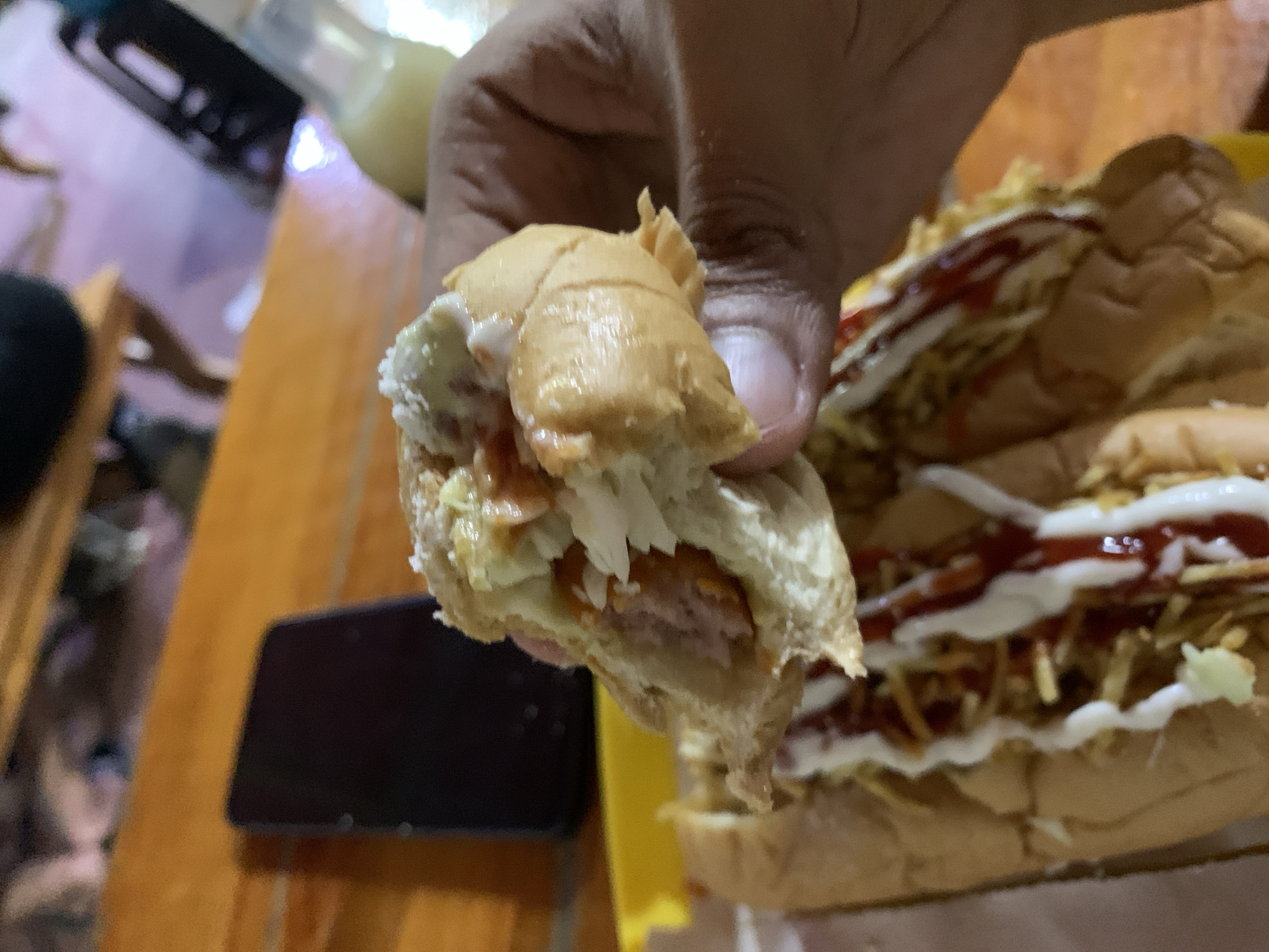 The image shows a close-up of a person's hand holding a large, open sandwich, likely a hot dog, with another similar sandwich visible in the background. The sandwich being held features a sausage, shredded white cabbage or coleslaw, and both white (possibly mayonnaise) and red (likely ketchup) sauces inside a toasted bun.
In the background, a second, fully assembled sandwich is visible, heavily garnished with shoestring potato chips ("papas rayadas"), ketchup, and white sauce, sitting on a yellow tray or plate. This style of hot dog with numerous toppings is characteristic of Venezuelan street food, known as "perros calientes."
The scene appears to be an indoor setting, possibly a casual eatery or home, with a wooden table serving as the surface. A dark-colored smartphone is partially visible on the table to the left. Overhead or artificial lighting illuminates the food. No discernible text is visible within the image. The location context for this food scene is Santa Ana, Venezuela.