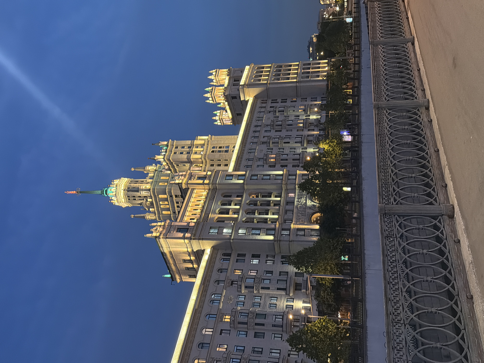 The image shows a grand, illuminated Stalinist skyscraper in Moscow, Russia, at dusk. The building is characterized by its symmetrical design, ornate architectural details, and multiple spires, one of which is topped with a flag. The windows of the building are lit, suggesting occupancy. In the foreground, a decorative metal fence borders a river or canal, with trees and streetlights visible along the embankment. The sky is a deep blue, transitioning into night, with a faint cloud streak. The overall scene conveys a sense of grandeur and urban beauty.