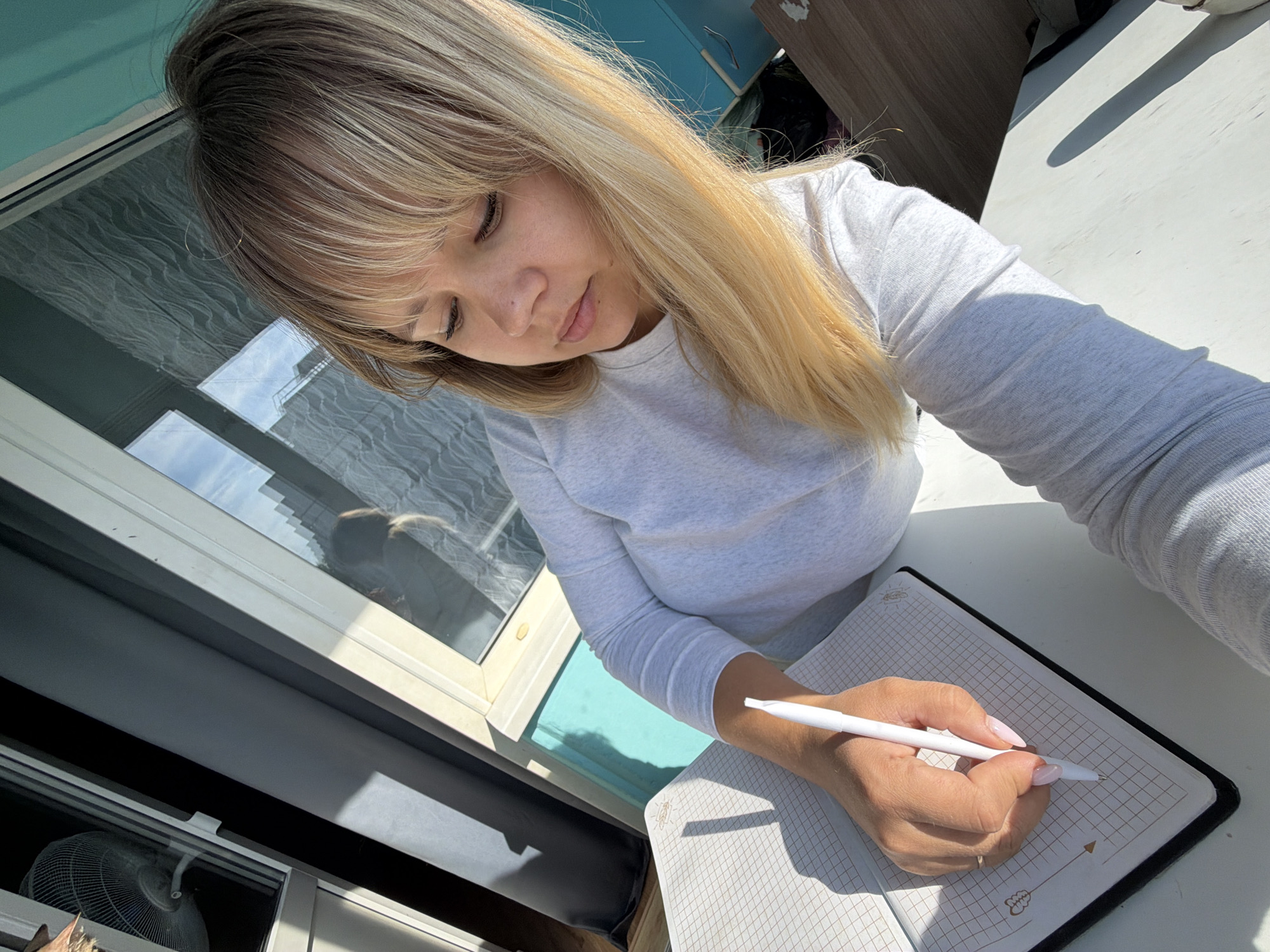 An overhead, close-up shot depicts a young woman engaged in writing or drawing indoors in Chaikovsky, Russia. Her blonde hair, styled with bangs, falls around her face as she leans over a grid-patterned notebook, eyes cast downwards in concentration. She is wearing a light grey long-sleeved shirt. Her right hand, featuring long light pink nails and a ring on her ring finger, holds a white pen poised over the page.
The scene is brightly lit by natural daylight, casting strong, distinct shadows on the white table where she works. To her left, a large window reflects the clear sky and outlines of distant buildings, suggesting a bright and clear weather day. A white fan is visible through the lower section of the window. The notebook shows a faint grid pattern and small decorative elements, including an arrow and what appears to be a stylized heart or seed motif, on the bottom right page. The overall impression is one of quiet focus and calm.