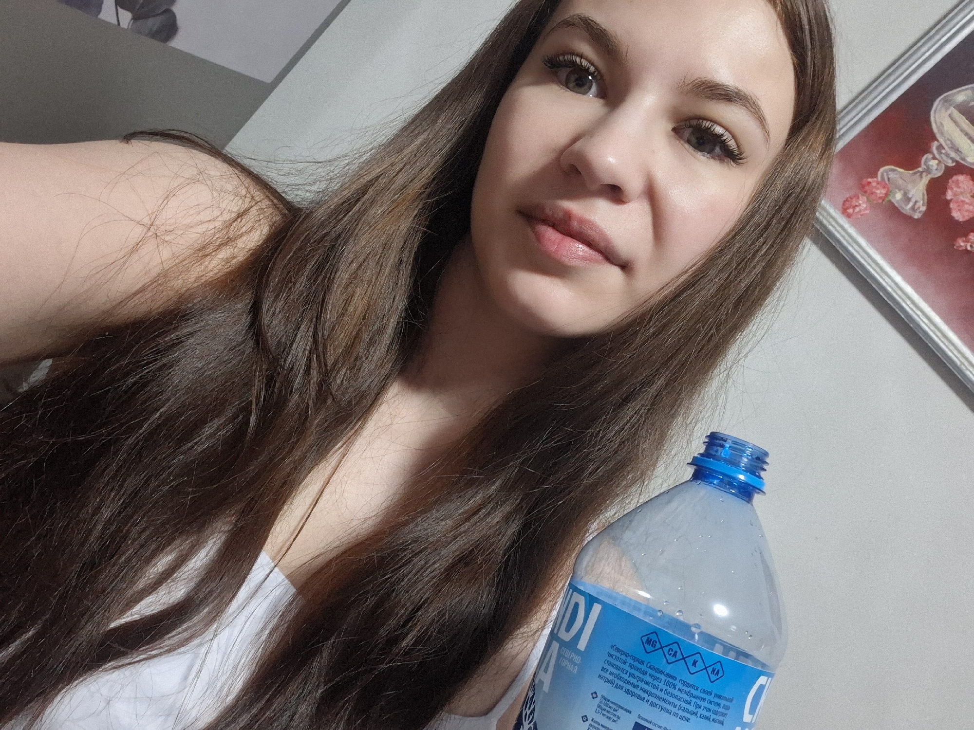 The image is a selfie of a young woman with long, brown hair. She is holding a blue plastic water bottle with Russian writing on the label, which includes text mentioning "Severno-gornaya Skandinavia" ("Northern Mountain Scandinavia") and its process of water purification, mentioning that it contains calcium, potassium, magnesium, and sodium. Based on the brand name, the location of the photograph might be considered near Chaikovsky, Russia, due to the brand "Северно-горная", which translates to "Northern Mountain". The woman wears a white top. In the background, there is a framed picture hanging on a light-colored wall. The lighting appears soft.