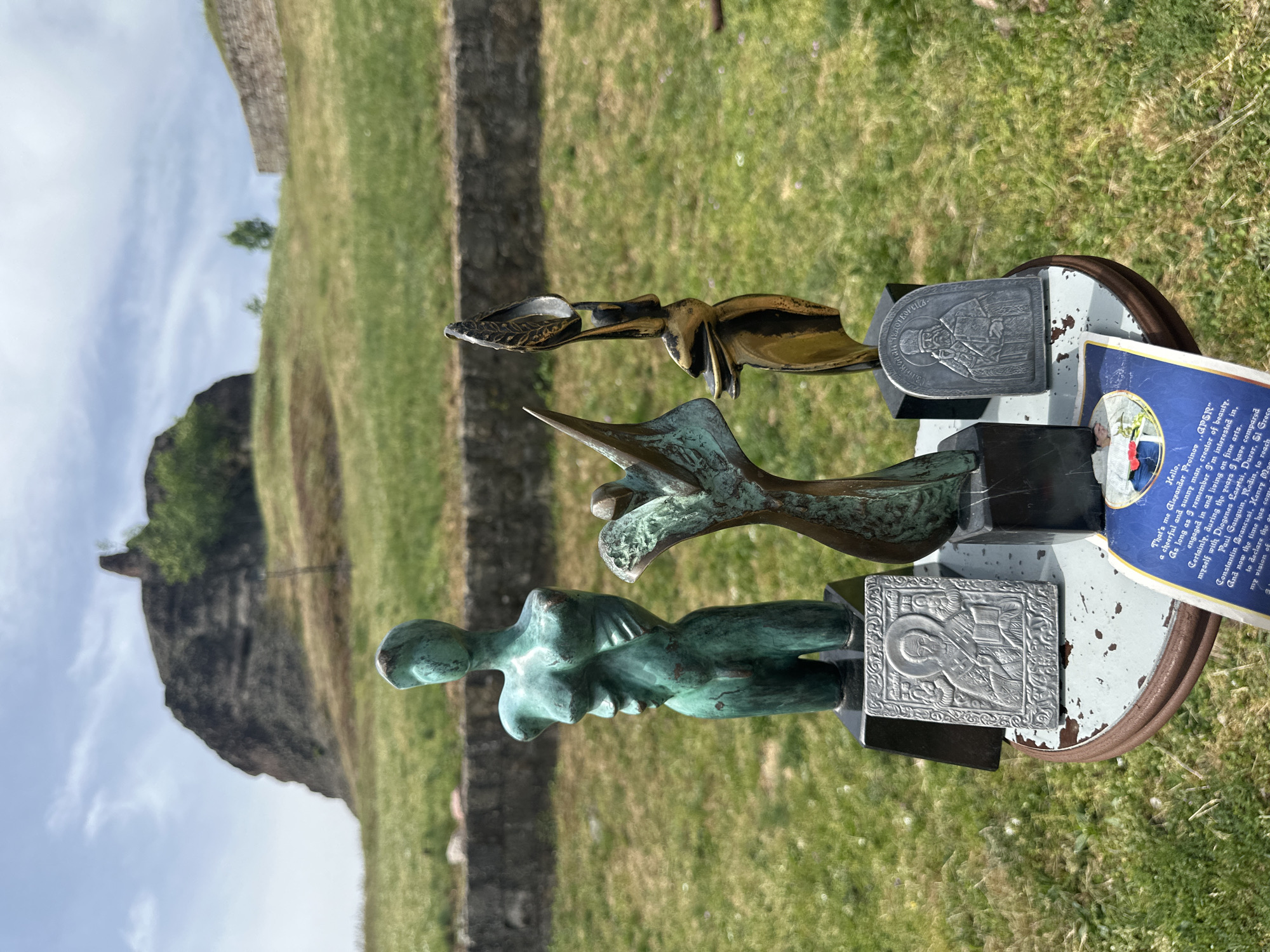 The image shows a collection of three metal sculptures on a pedestal, set against a backdrop of a grassy landscape and a rock formation in Belogradchik, Bulgaria. The bottom sculpture is a verdigris-colored figure of a woman's torso. Above it is a green and bronze sculpture that looks like a stylized bird or dragon. The top sculpture is a more ornate golden piece. Each sculpture has a grey metal plaque attached, with religious iconography. Beside the sculpture is a small blue information sign. The background features green grass and a rock formation, characteristic of the Belogradchik Rocks area.