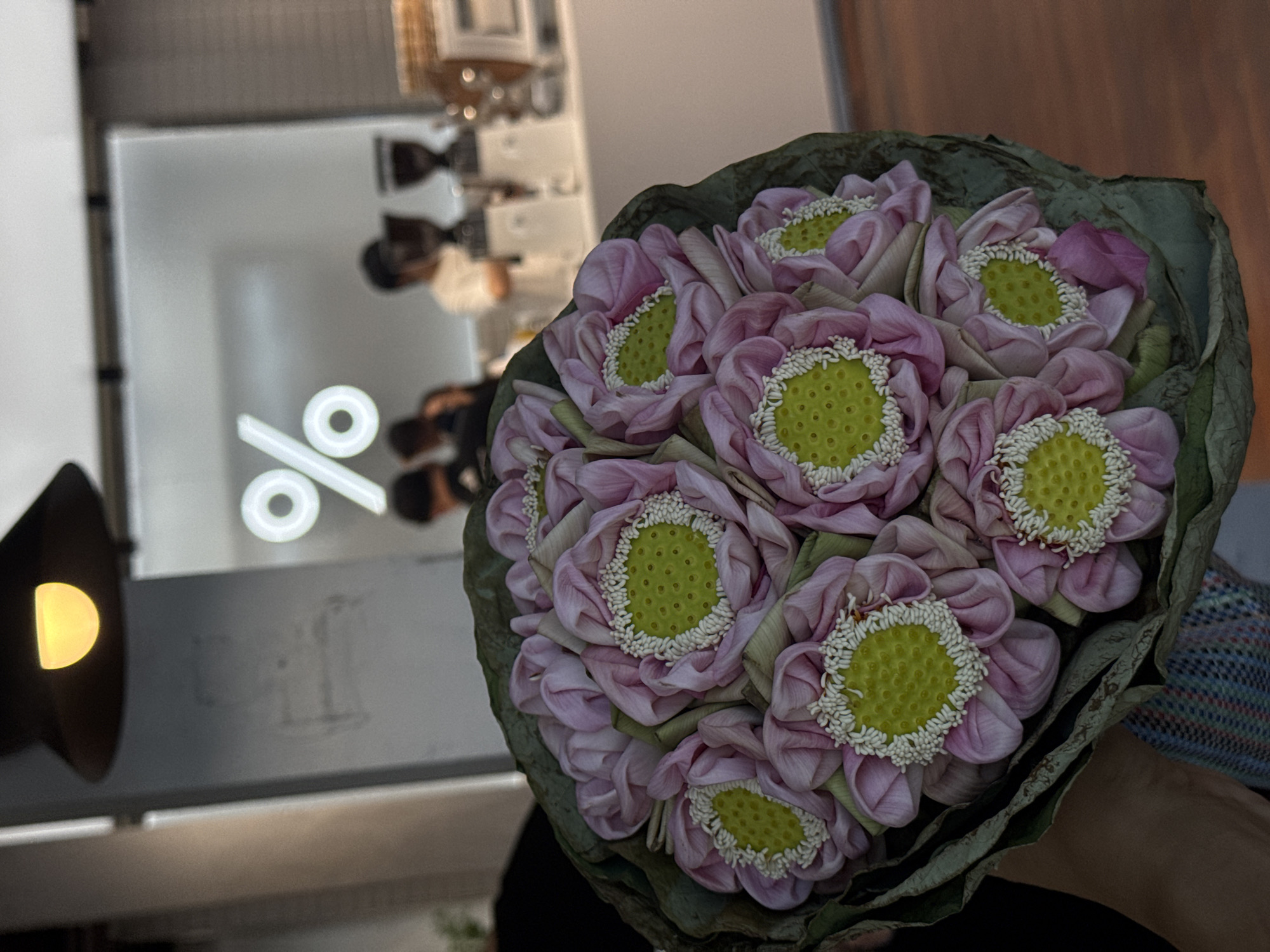 A bouquet of pink lotus flowers with green seed pods and white stamen is held in a person's hand. In the background, a white wall displays a large, illuminated percentage sign (%). Several people are blurred in the background, some appearing to be standing behind a counter in a cafe. Coffee machines and cups are visible on the counter. The scene suggests a cafe setting, possibly in Bangkok, Thailand, given the prevalence of lotus flowers in Thai culture and the distinctive "%" logo often associated with certain coffee chains. The lighting indicates it is likely evening or indoors with artificial light.
