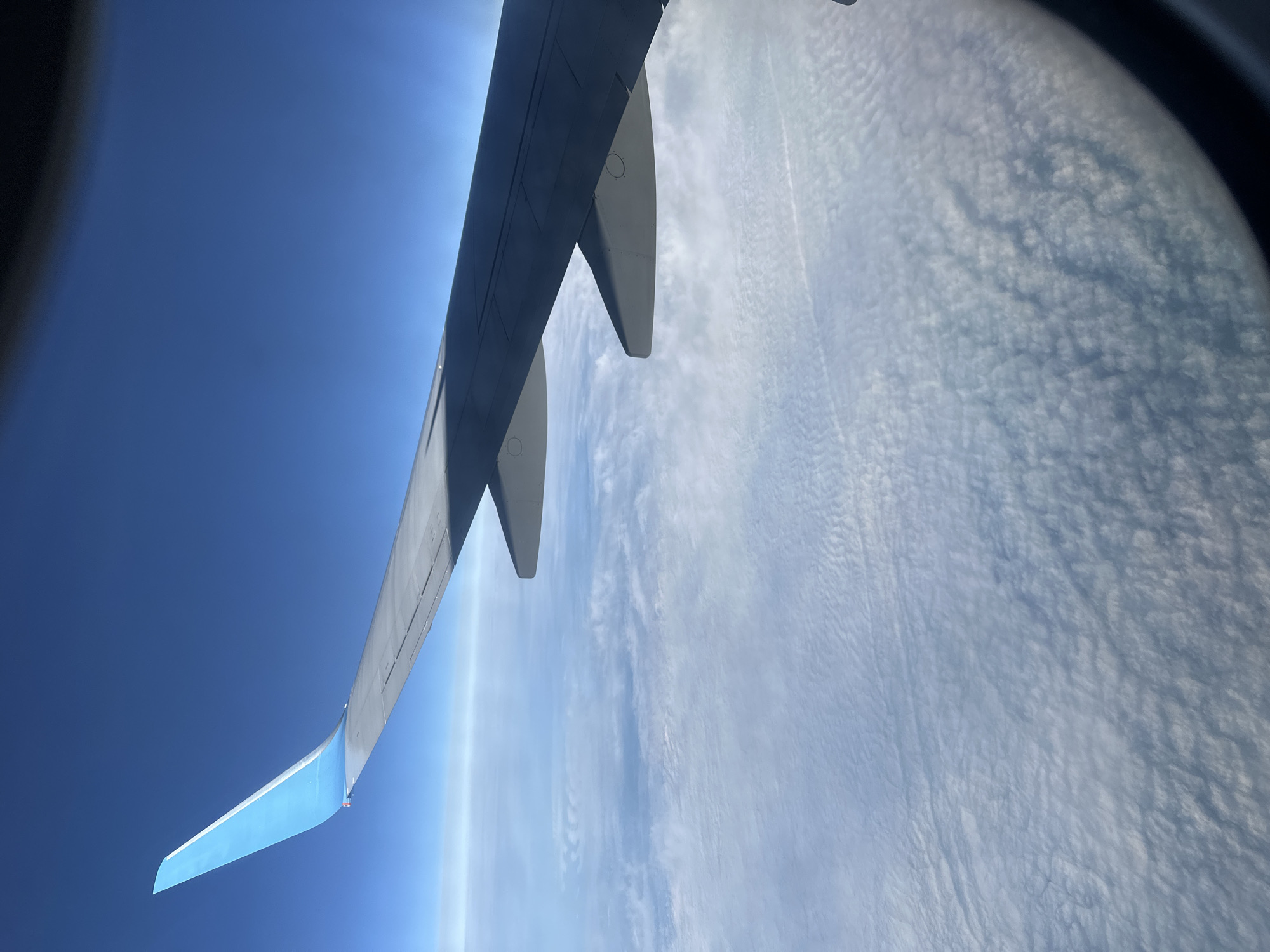 The image shows an airplane wing seen through an airplane window. The sky is visible above the wing and a layer of clouds is below the wing. There is no indication of Kitsjanovo, Russia, or any people, subjects, objects, or actions besides the wing, clouds, and sky seen from the airplane window.
