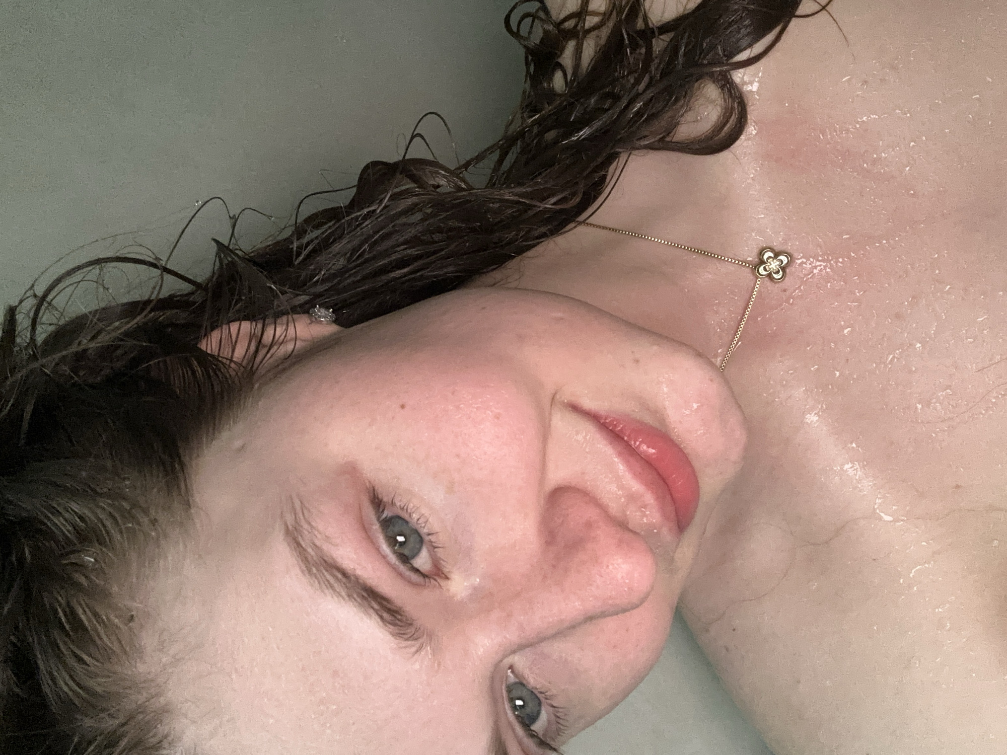 A close-up, upside-down view of a fair-skinned person with wet, dark brown hair. The person has grey-blue eyes, light pink lips, and freckles. A gold necklace with a flower-shaped pendant is visible on their neck. Droplets of water are present on their skin and hair. The background appears to be a solid light grey or green color. There is no information to suggest the location is Kazan, Russia.