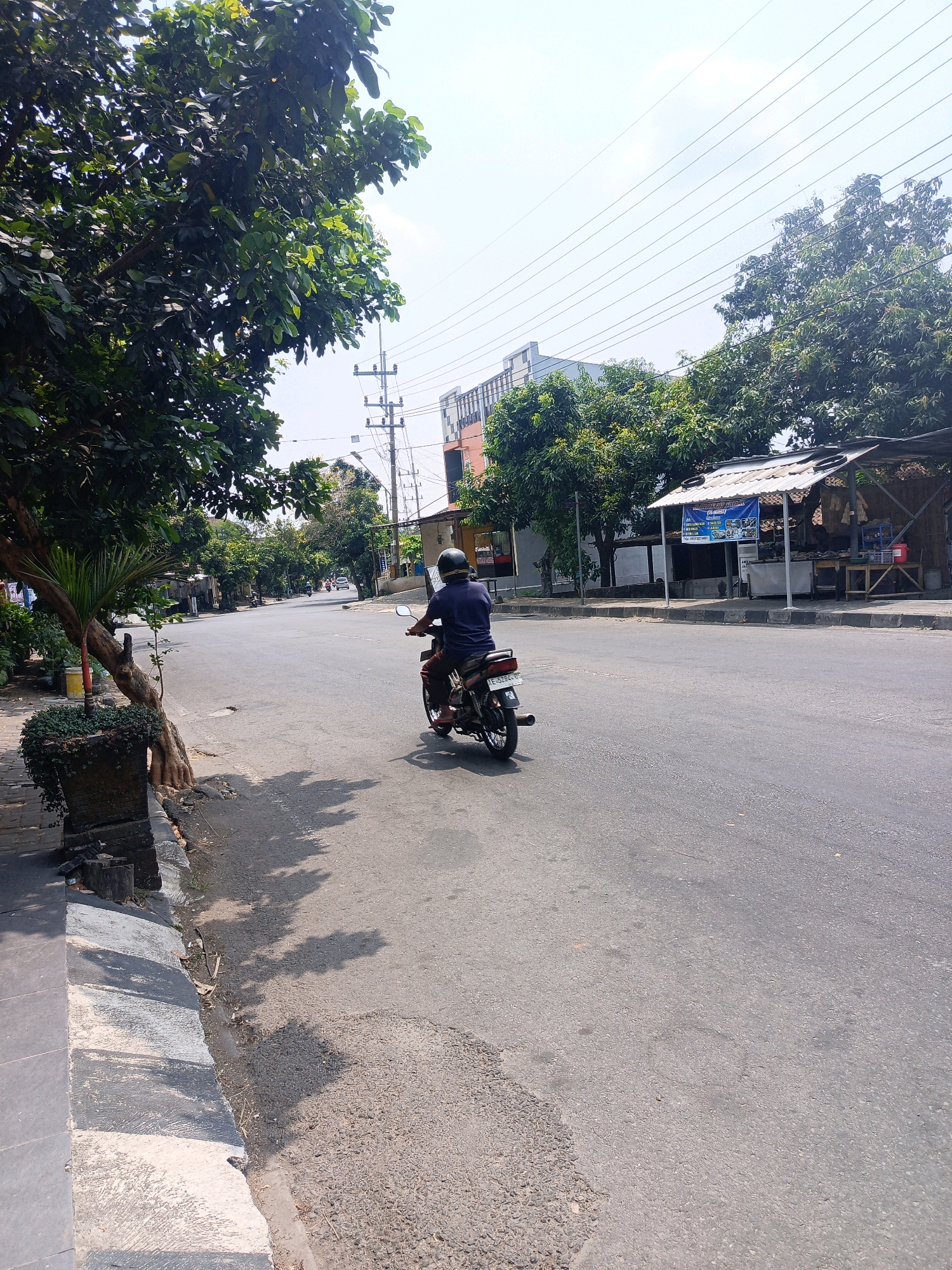 The image depicts a street scene in Mangkujayan, Indonesia, under a clear, bright sky. A lone individual is riding a motorcycle on the right side of the asphalt road, moving away from the viewer. The rider is wearing a dark helmet and a dark blue shirt. The motorcycle is black with red accents and a license plate that appears to read "E 5284 US".
To the left of the road, a sidewalk with a textured surface is visible, bordered by a curb. A potted plant with cascading greenery sits beside a tree with a rough, brown trunk and dark green leaves. Further to the left, more trees line the sidewalk.
The road itself is paved and appears to be in moderate condition, with some patches and wear visible. In the background, across the street, there are several buildings and structures. One building is a multi-story structure with a light-colored facade. Adjacent to it is a small open-air stall or shop with a corrugated metal roof, displaying a blue banner with text and images. A few trees are scattered throughout the background. Power lines are strung overhead, supported by utility poles. The overall environment suggests a moderately developed urban or semi-urban area.