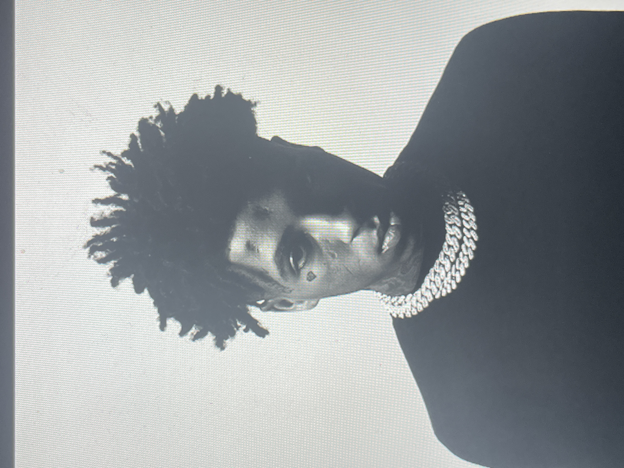 A portrait photograph captures a young man in what appears to be a studio setting, predominantly in black and white or a highly desaturated grayscale. The image is rotated 90 degrees clockwise, showing the subject's head tilted to the right.
The central subject is a man with dark, textured hair, possibly dreadlocks or a similar style, styled to appear spiky or unkempt. He has distinct facial features, including a small tattoo shaped like a cross beneath one eye and potentially another faint mark on his cheek or forehead. His expression is serious or stern, and he appears to be looking directly or slightly away from the camera.
He is wearing a dark-colored garment, likely a shirt or jacket, and a prominent, heavy, silver-colored Cuban link chain around his neck. The chain's links are highly reflective, catching light and standing out against his dark clothing.
The background is a plain, light-colored surface, possibly white or a very light grey, which creates a strong contrast with the dark tones of the subject and his clothing. The overall image quality shows a pixelated grid pattern, suggesting it is a photograph taken of a digital image displayed on a screen or monitor rather than an original print. There are no visible location cues within the image itself, nor is any text discernable. The activity depicted is a posed portrait.